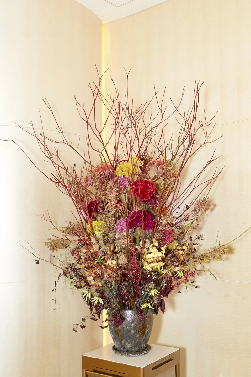 Tall sculptural floral arrangement with red branches, celosia, orchids, and mixed textures in a bronze vase for a hotel lobby corner.