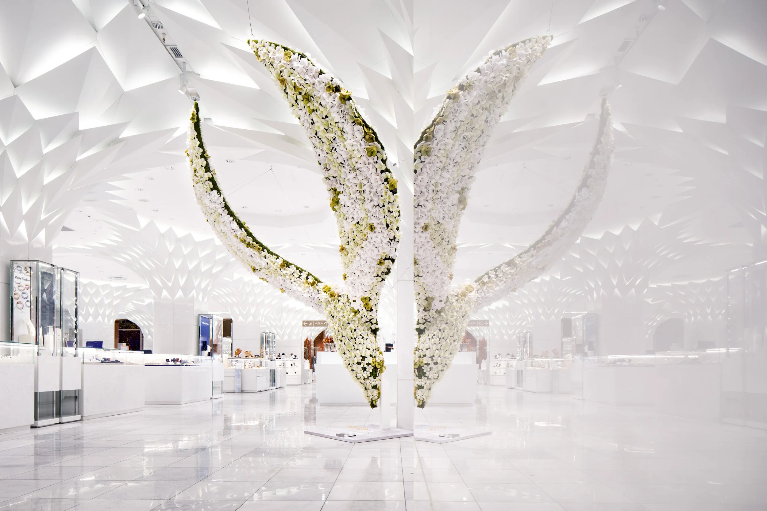White indoor space with a large, symmetrical flower arrangement in the shape of a butterfly or a heart, made of white flowers with some green accents, hanging from a white ceiling with geometric patterns.