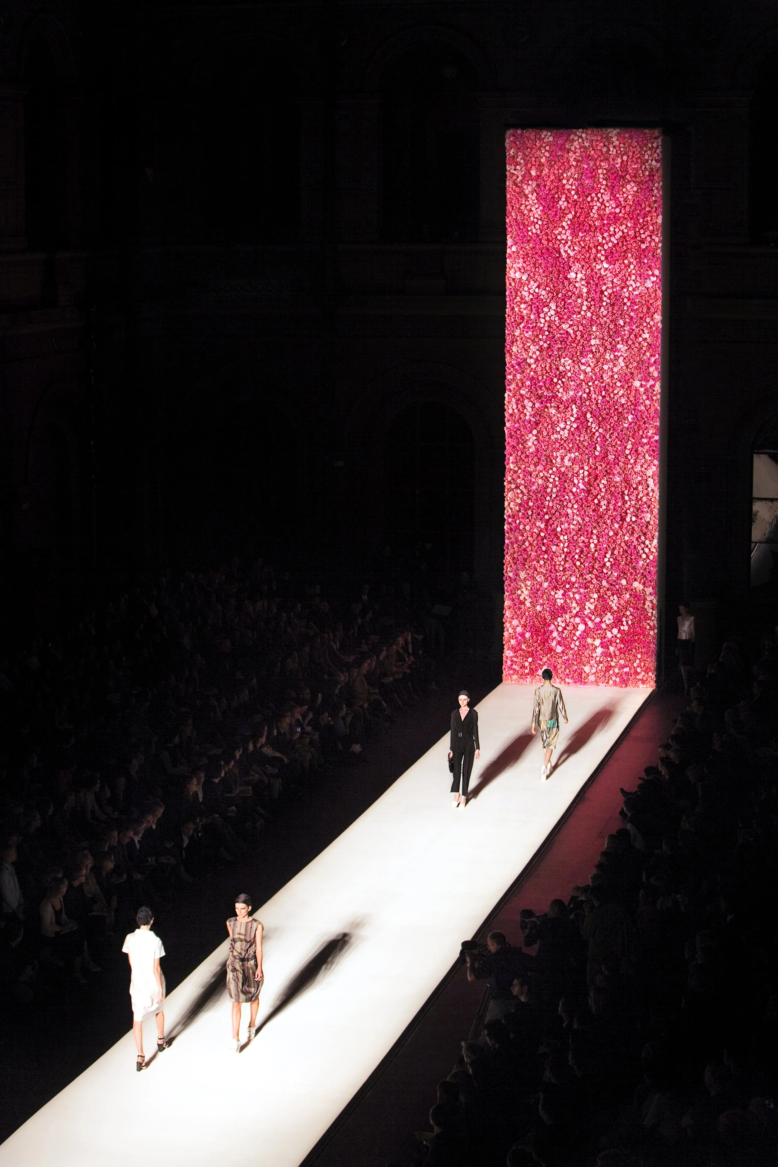 Models walk down runway during a fashion show, with a large pink floral backdrop and seated audience on both sides.