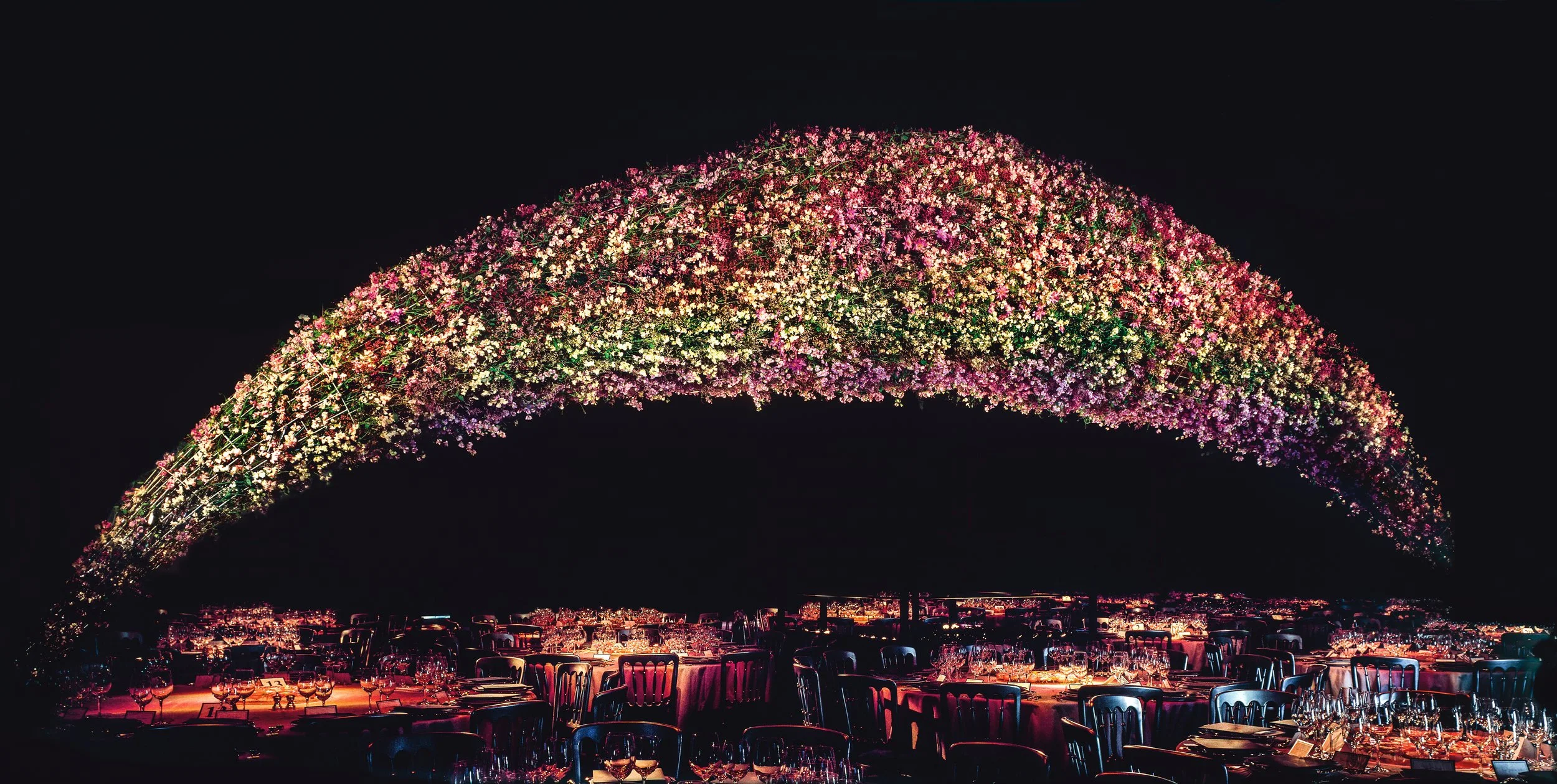 Luxury corporate banquet with vast rainbow arch of thousands of flowers suspended above tables, Daniel Ost signature floral artistry.
