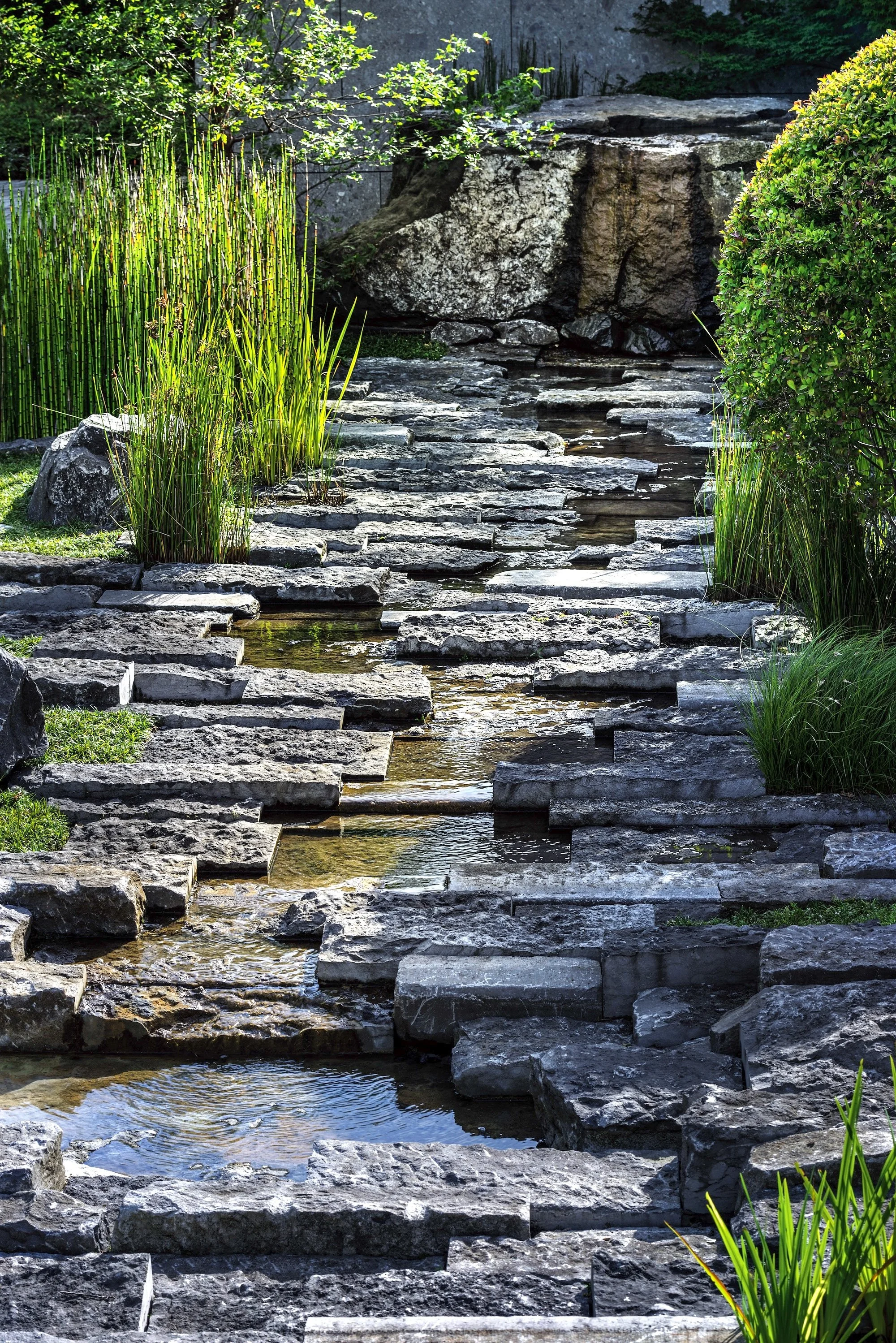 Naturalistic stone water feature with layered rock steps, flowing stream, and vertical grasses in a private garden.
