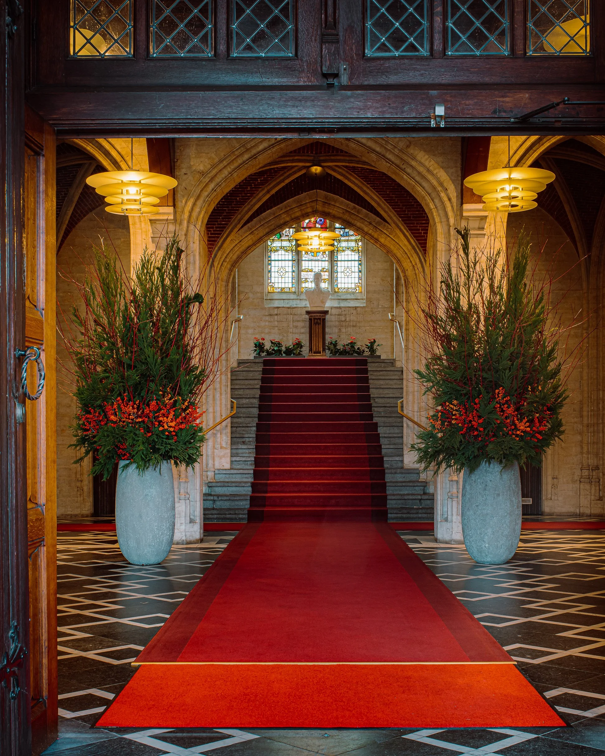 Elegant corporate floral decoration with tall red and green vases framing the entrance of a Gothic hall with red carpet and stained-glass windows.