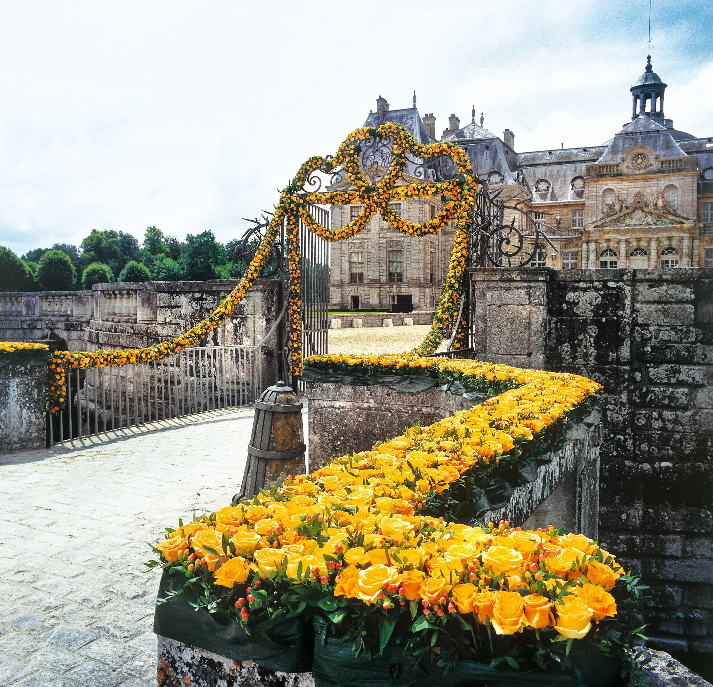 Castle entrance decorated with garlands of vibrant yellow roses and orange accents, framing ornate wrought-iron gates for a regal wedding arrival.