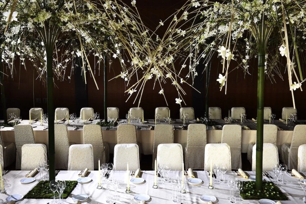 Luxury wedding dining setup with towering bamboo centerpieces adorned with cascading orchids and grasses, creating an intimate garden-like atmosphere indoors.
