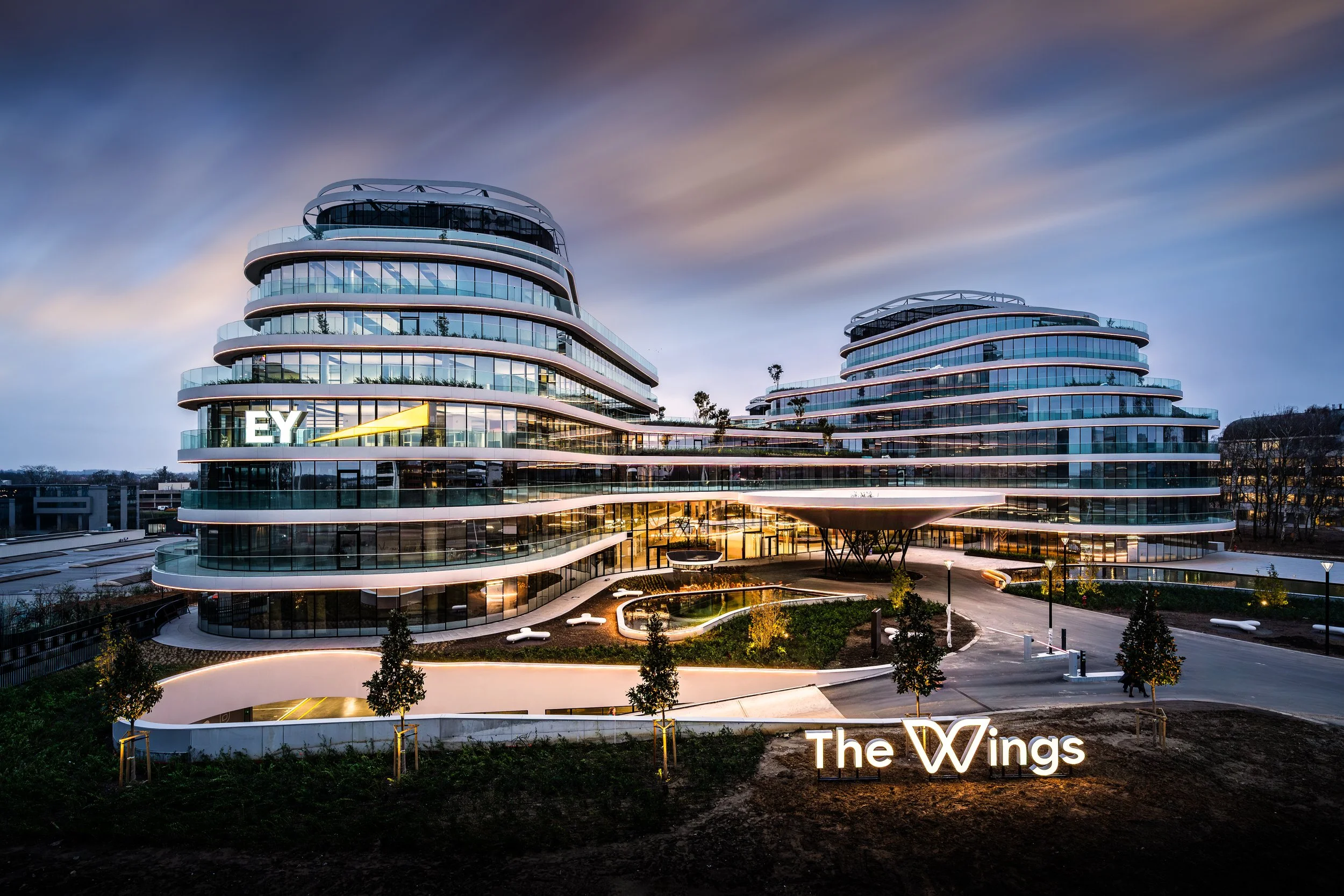 Contemporary glass office complex with curved architecture, branded EY signage, landscaped forecourt, and illuminated façade.
