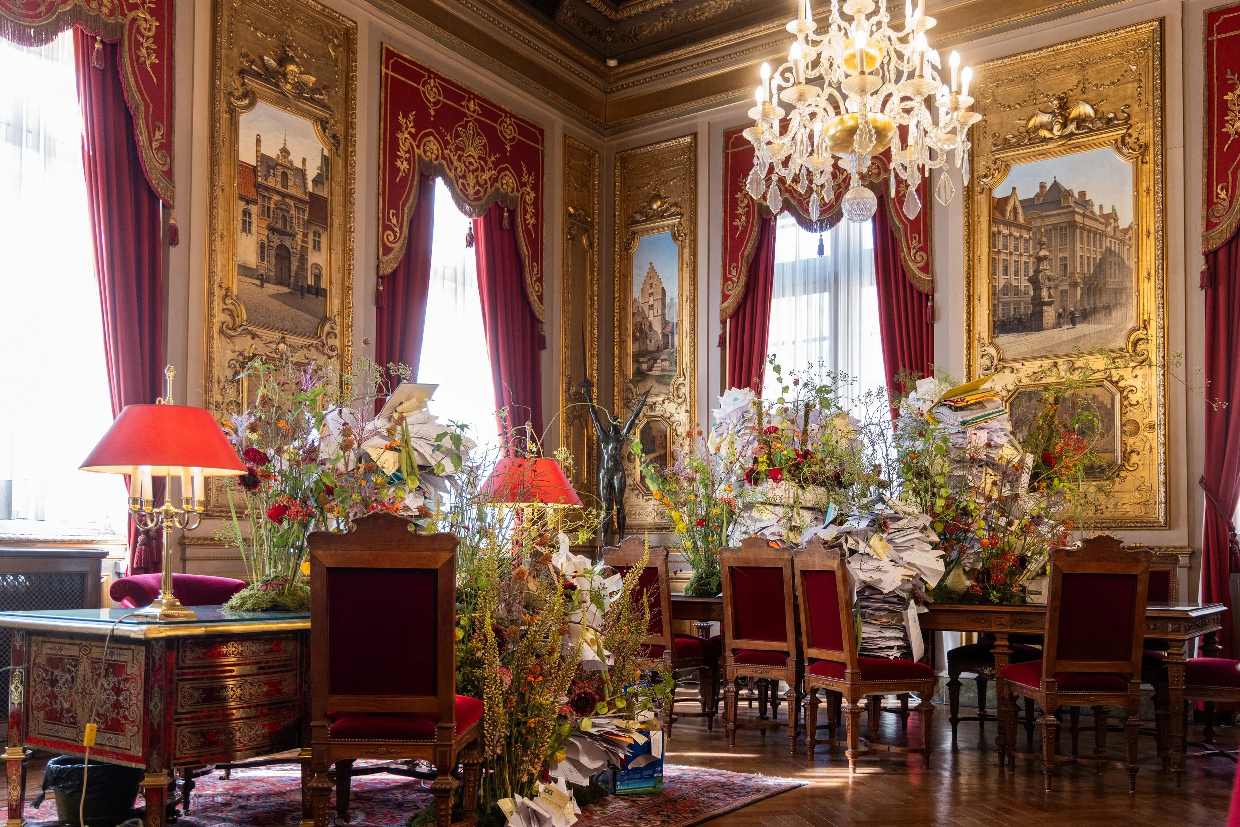 Opulent floral arrangements with paper art integrated into historic golden interior of a ceremonial hall with red velvet curtains and chandeliers.