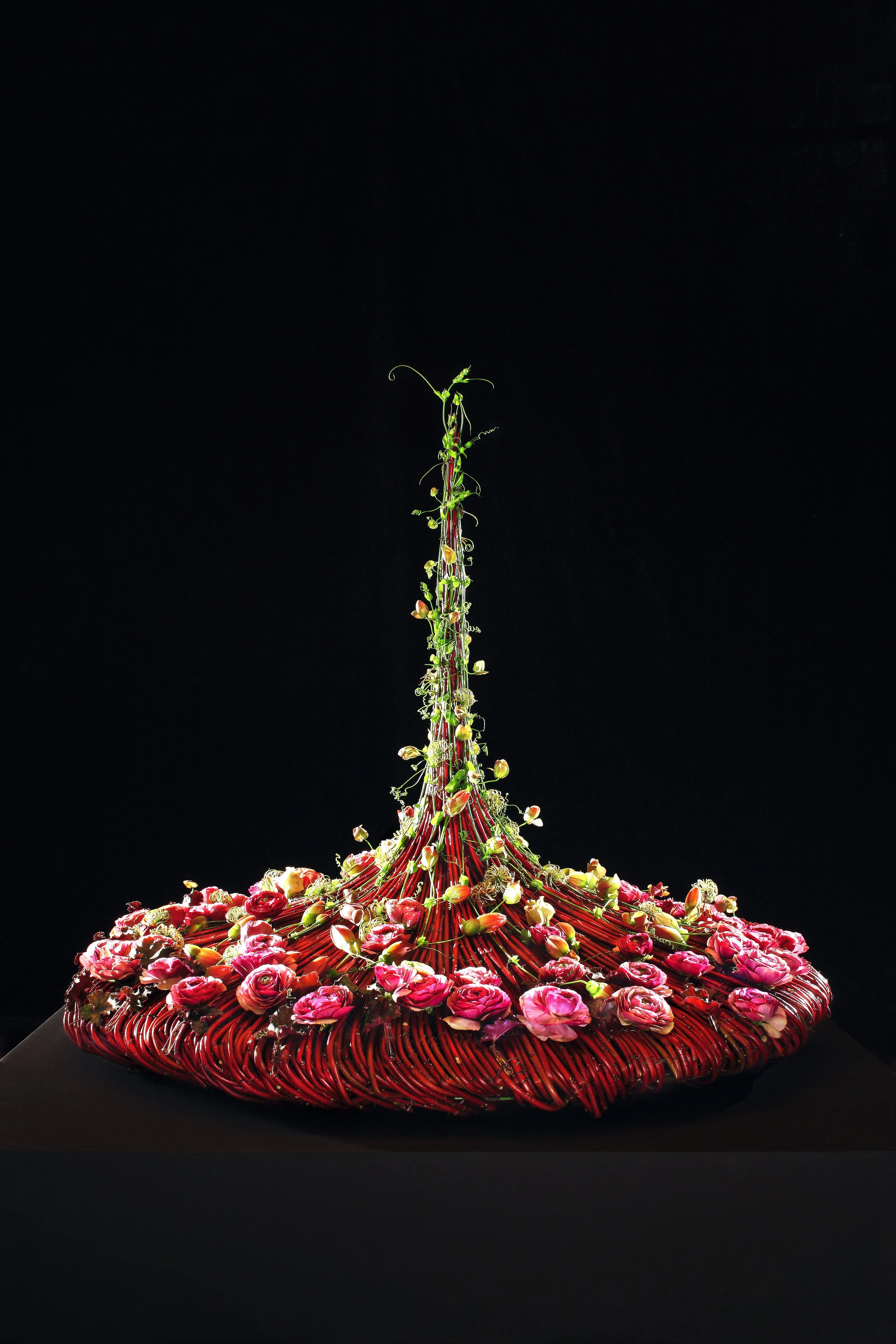 A sculpture resembling a fountain made of woven red material with pink flowers and green plants on top, against a black background.