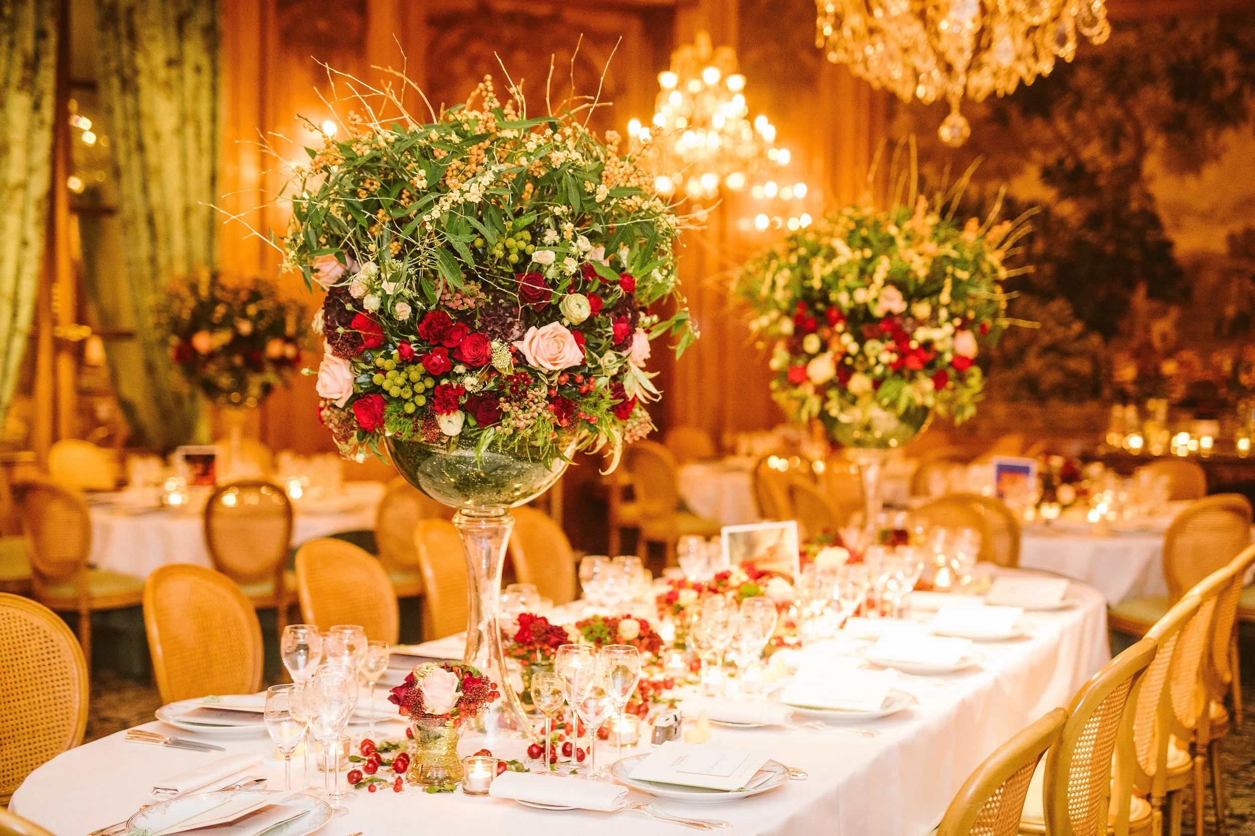 Luxury wedding table centerpiece with lush red and blush roses in tall glass vases, styled in a golden ballroom with chandeliers – elegant floral design by Daniel Ost.