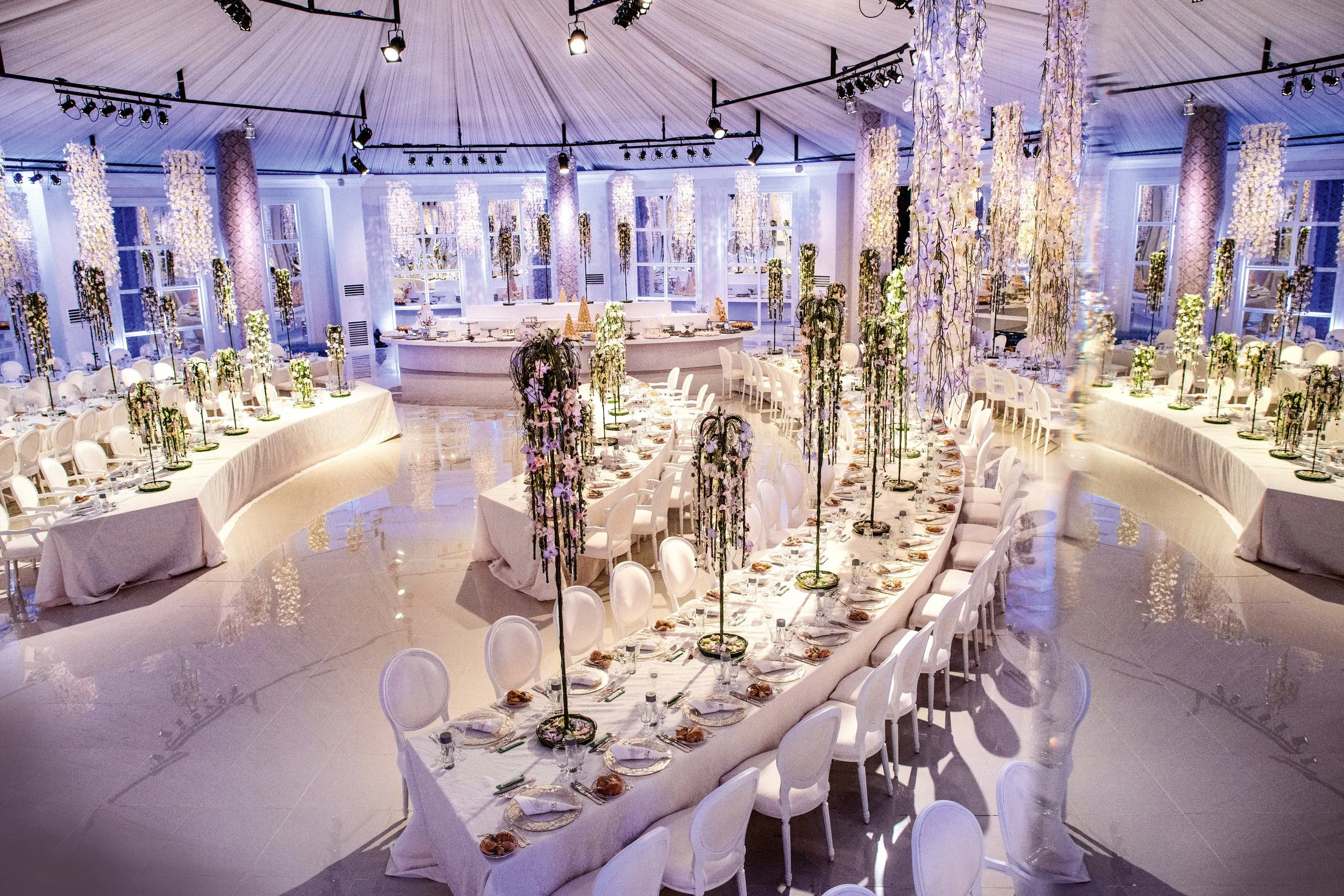 Modern wedding banquet with long curved tables, white draped seating, and tall cascading floral centerpieces in a luminous white and pastel setting.