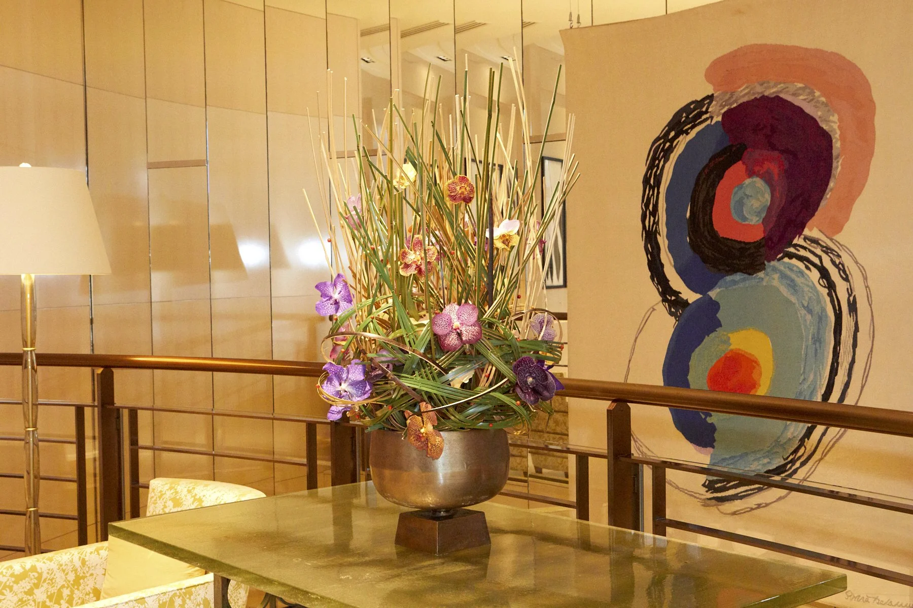 Hotel lounge floral arrangement with orchids and tropical leaves in a bronze vase, styled next to modern art décor.