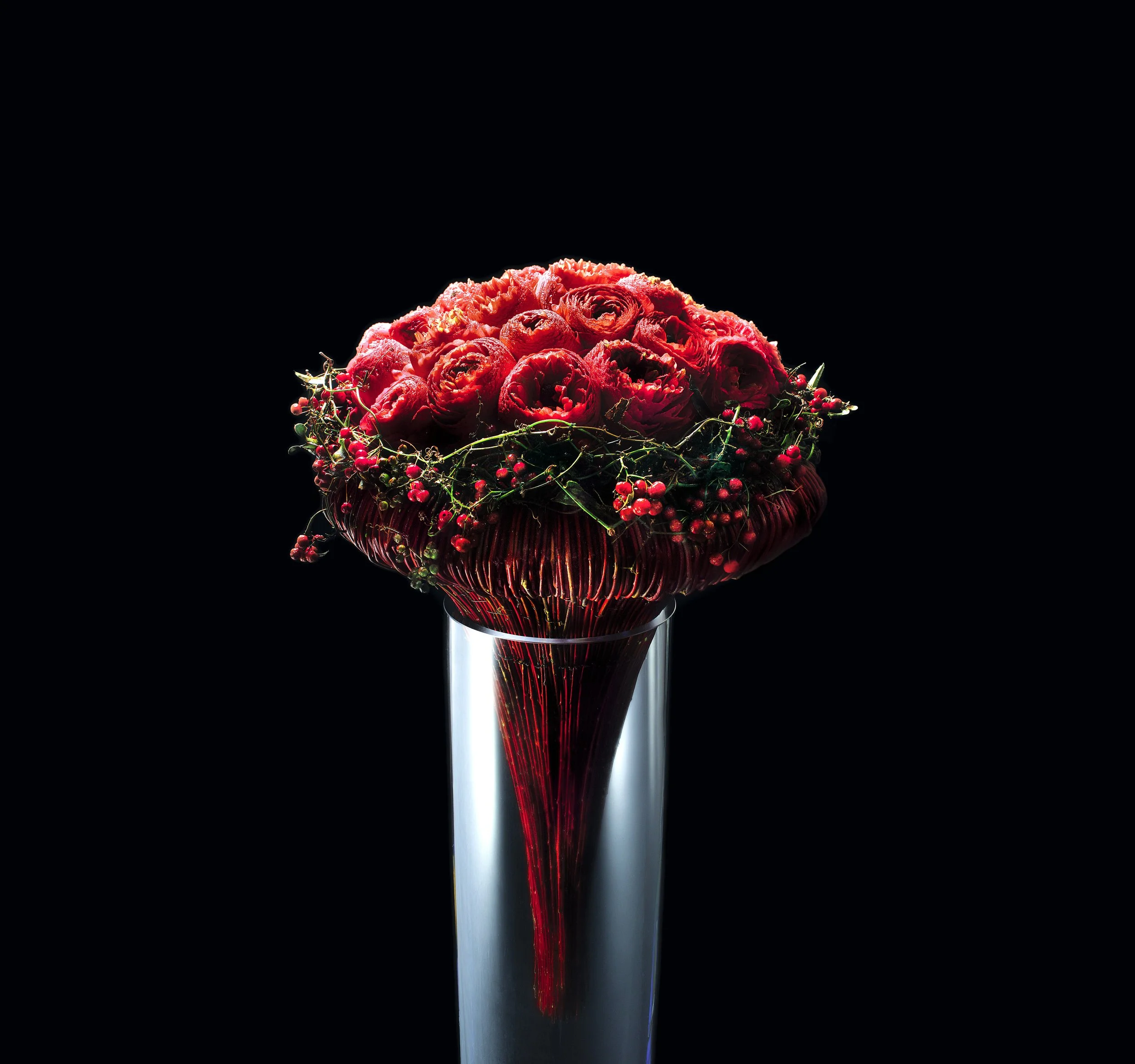 Sculptural bouquet of deep red ranunculus with berries, arranged in a structured red base on a tall glass pedestal.