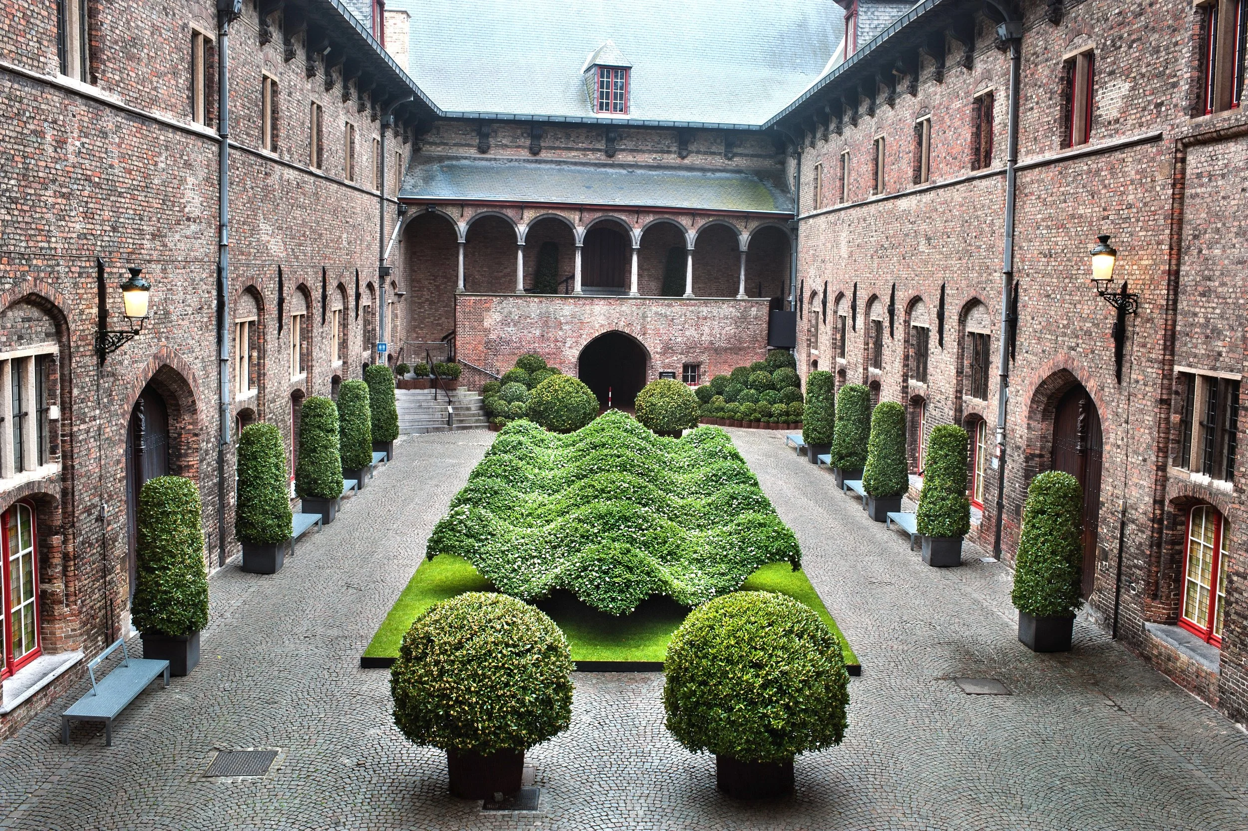 Historic courtyard garden with geometric topiary and undulating green sculptures surrounded by brick gothic architecture.