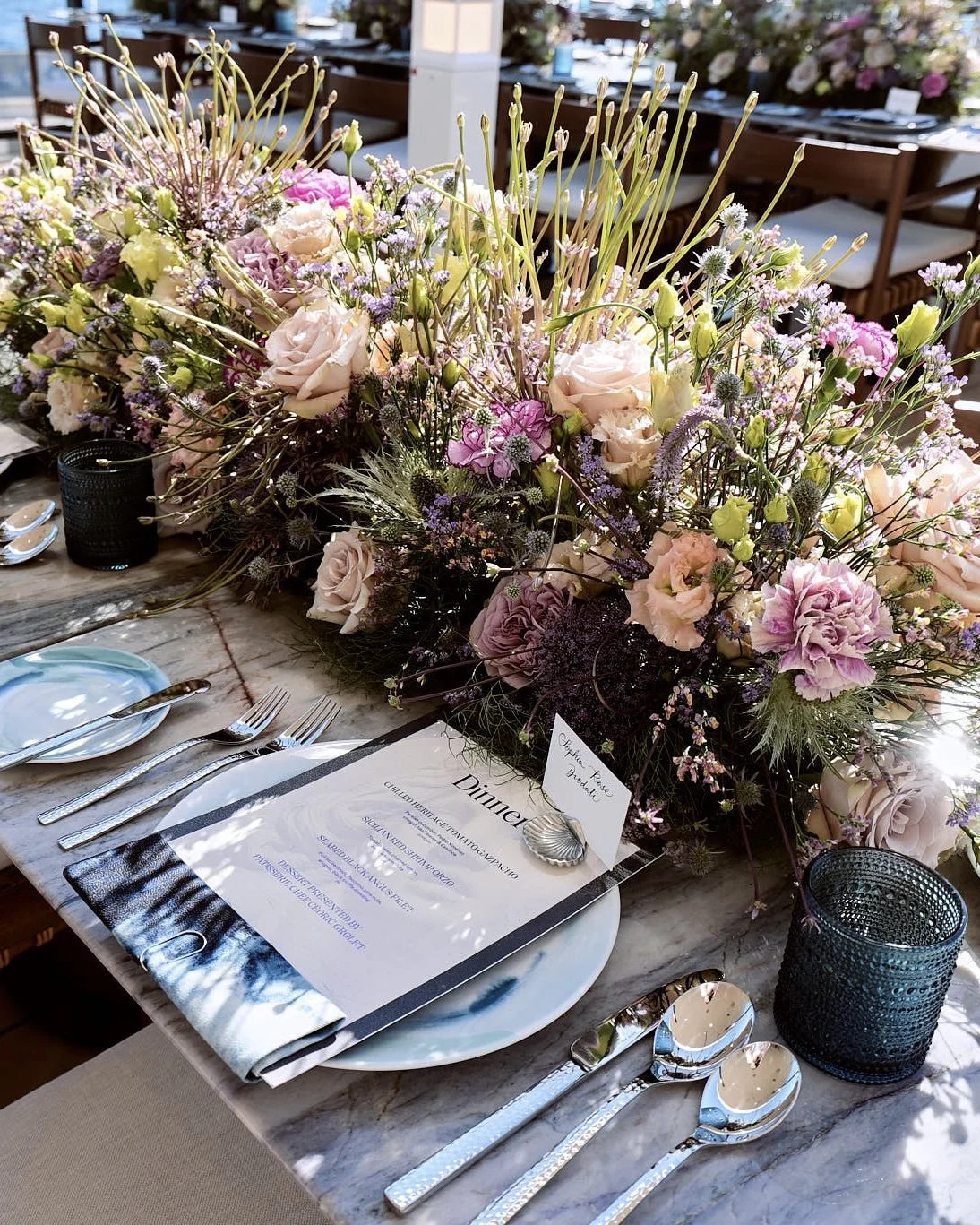 A table decorated with a large pastel-colored floral centerpiece, including roses, carnations, and other flowers. There is a dinner menu, silverware, and glassware on the table, with two black votive candle holders and a small place card that reads '