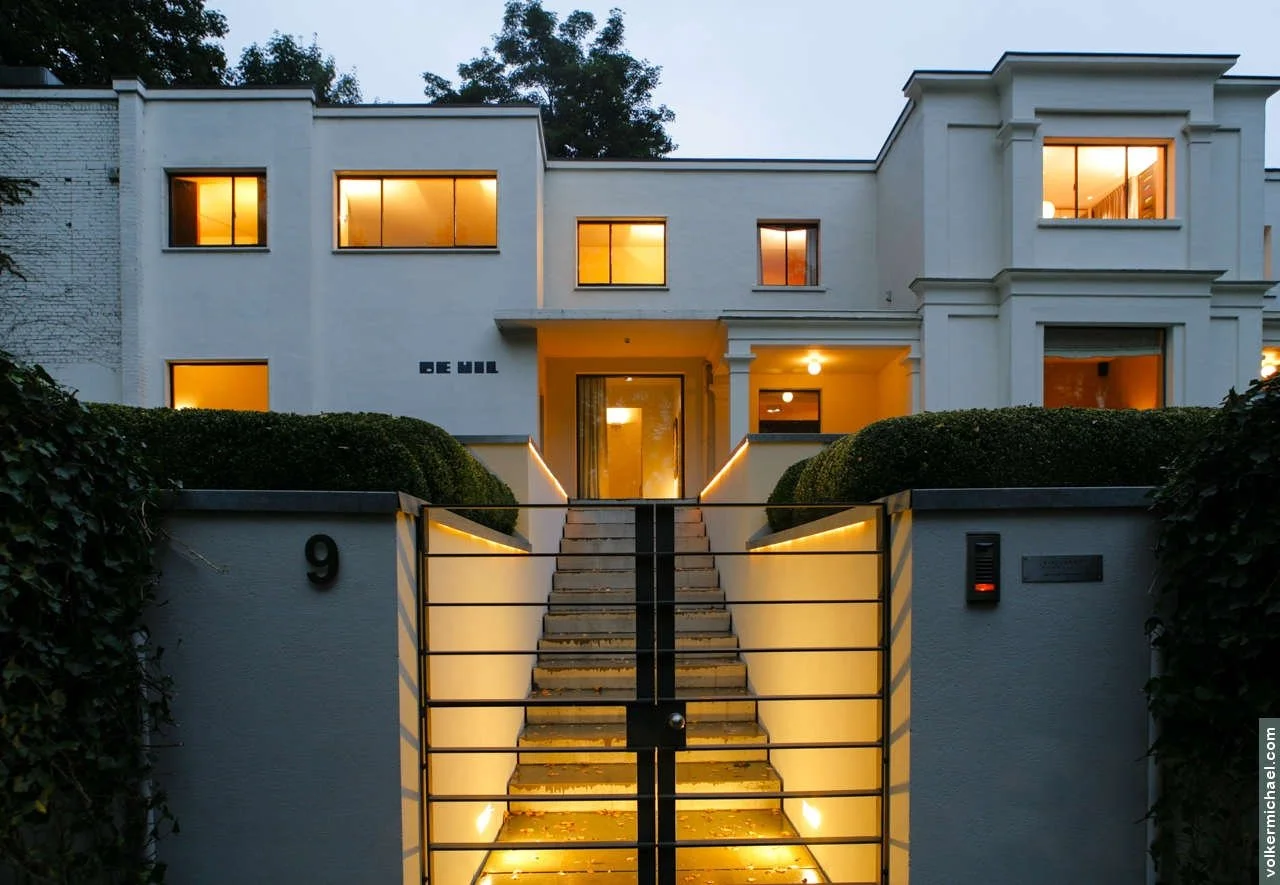 Contemporary private villa entrance with illuminated stairway, minimalist landscaping, and symmetrical trimmed hedges.