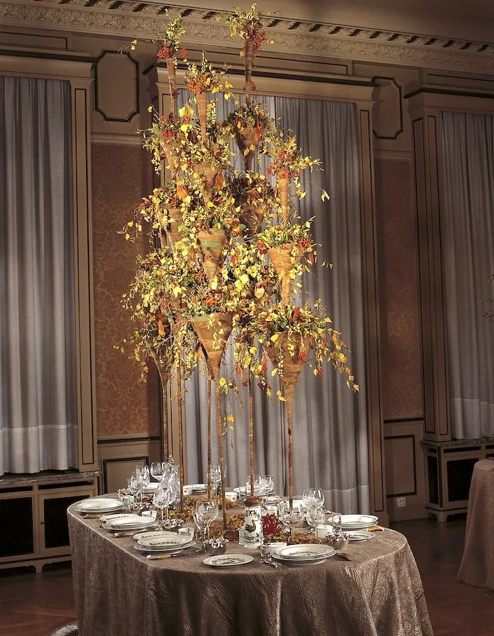 Tall table arrangement with golden foliage and orange accents in tiered cone vases, bringing warmth and seasonal charm to an intimate wedding dinner.