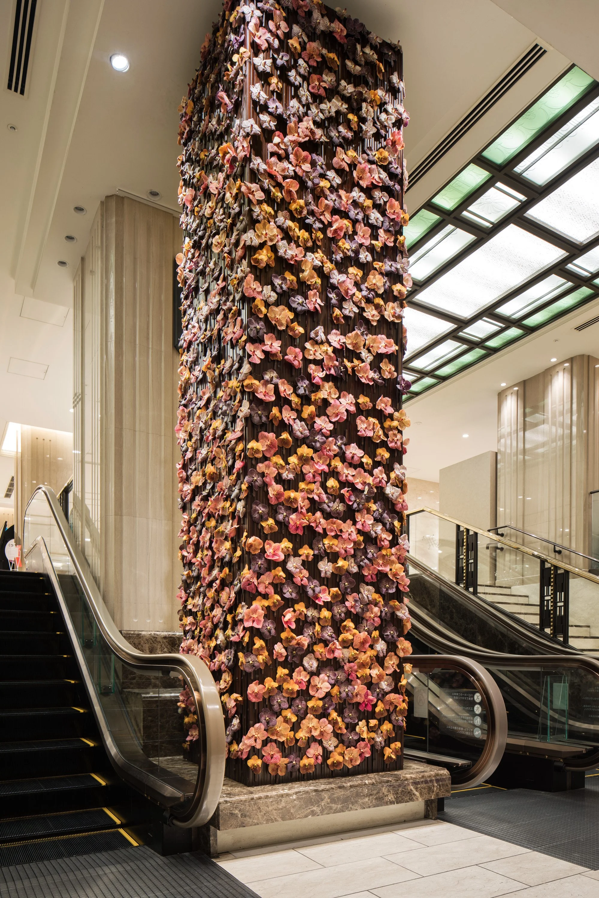 Decorative floral installation with pink, purple, and orange flowers on a dark vertical column inside a hotel lobby, with escalators and marble accents.