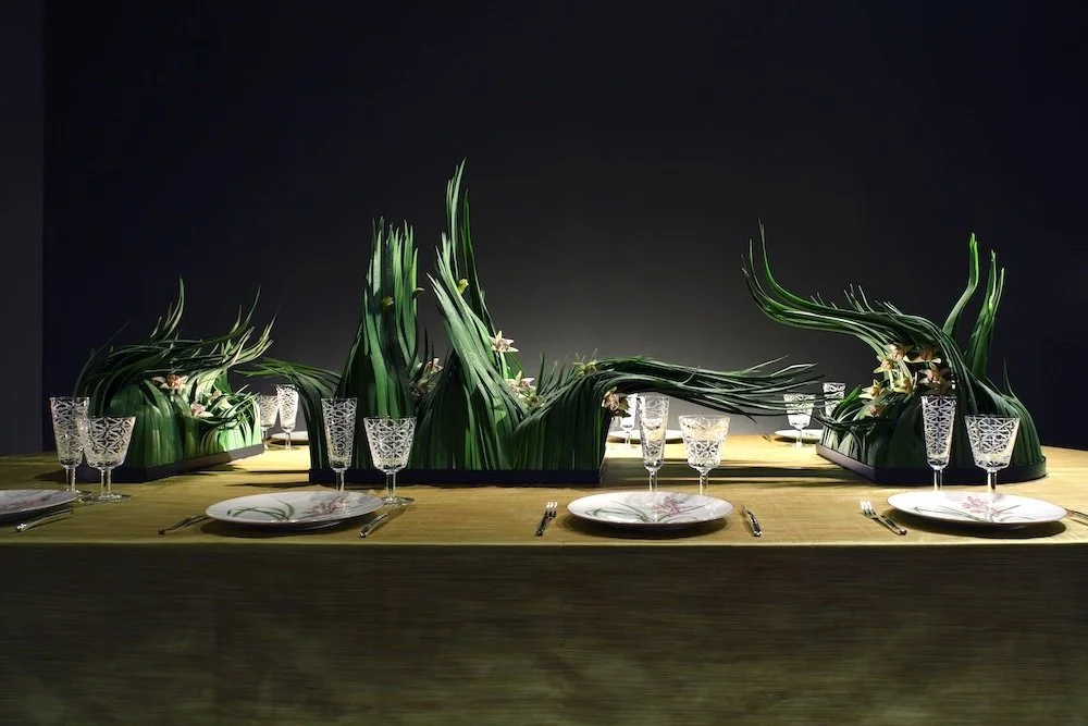 Unique wedding centerpiece with sweeping green leaves shaped into waves, accented with delicate flowers, perfect for a contemporary intimate dining setting.