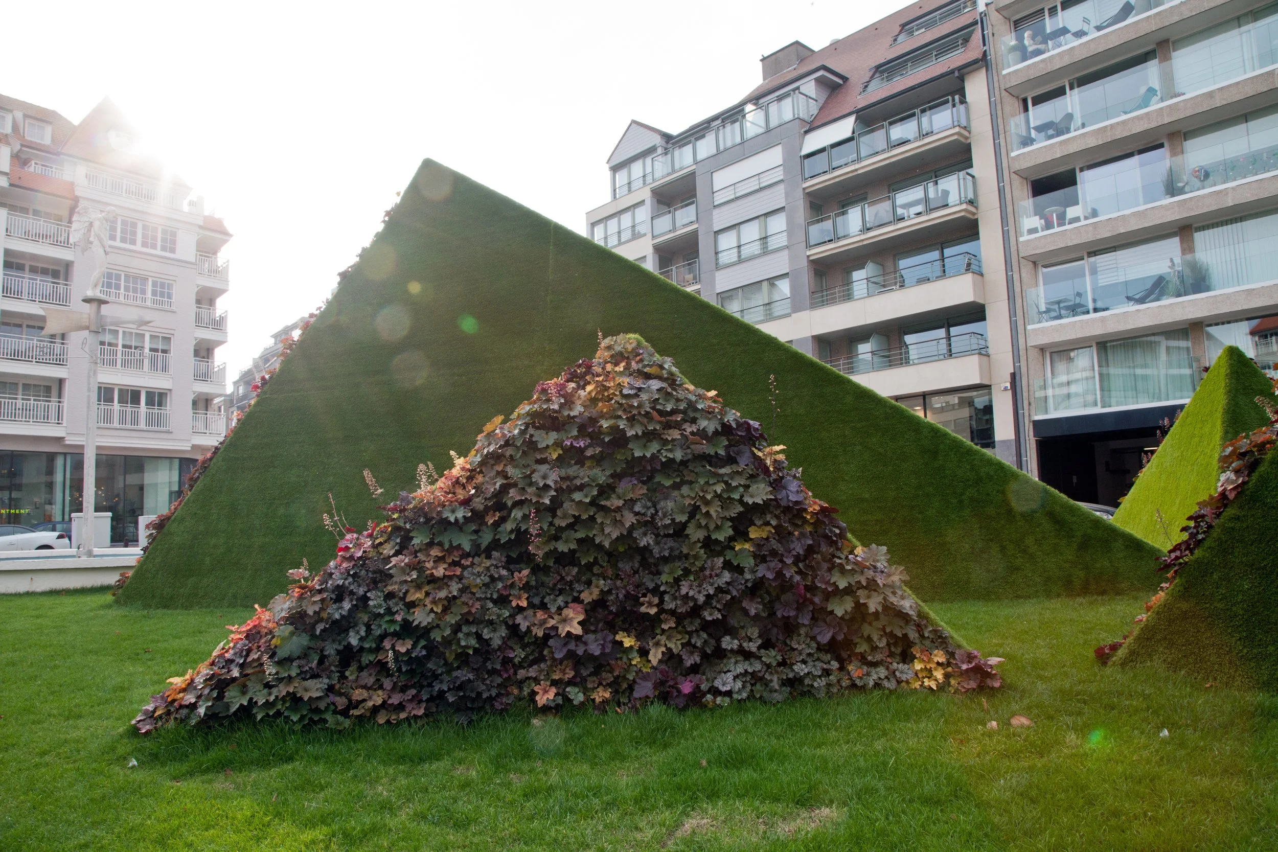 Contemporary landscape installation with grass pyramids and plant-covered geometric sculptures in a residential square.