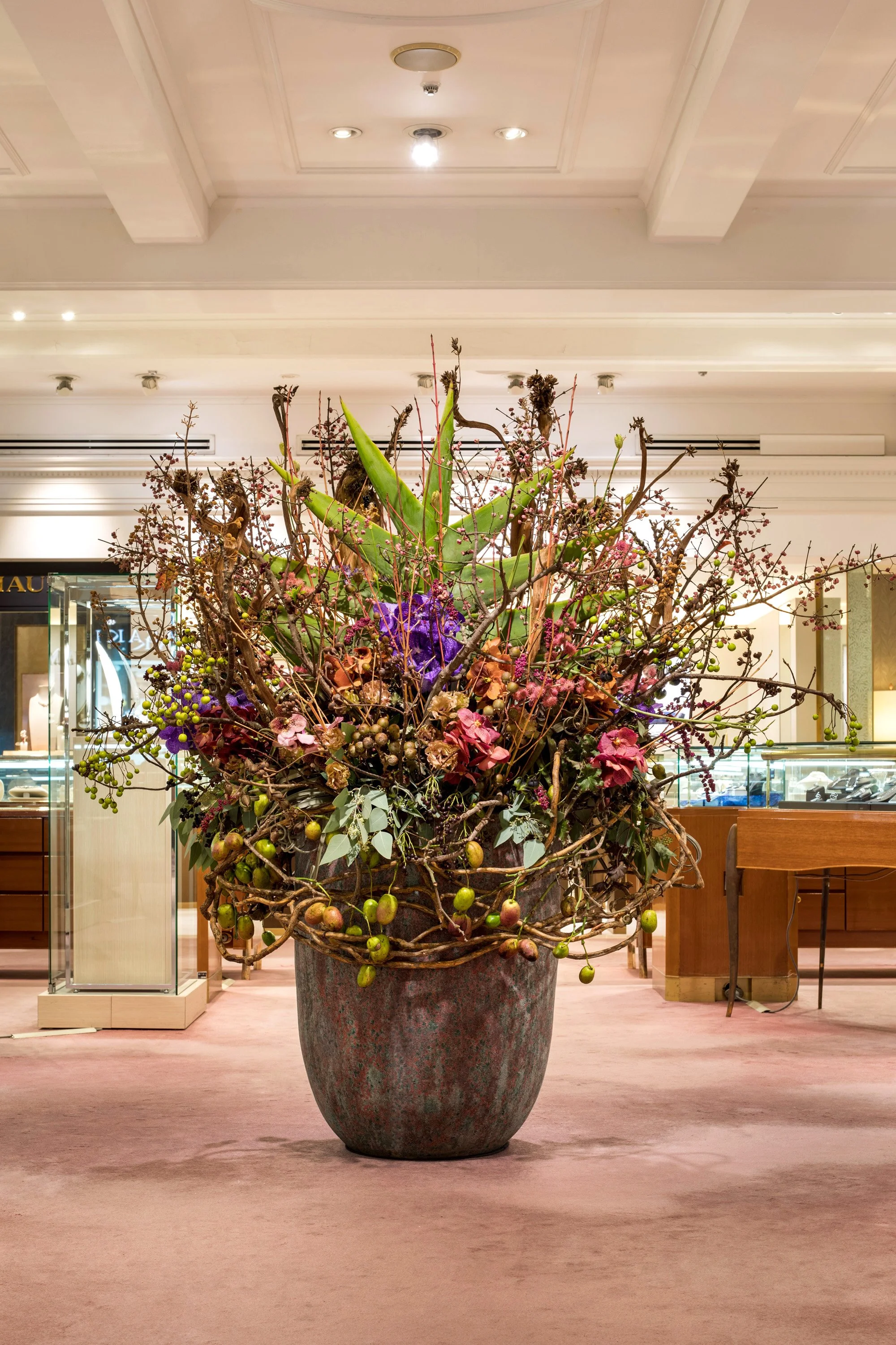 Large decorative floral arrangement in an indoor lobby setting with a pink carpet, illuminated ceiling, and display cases in the background.