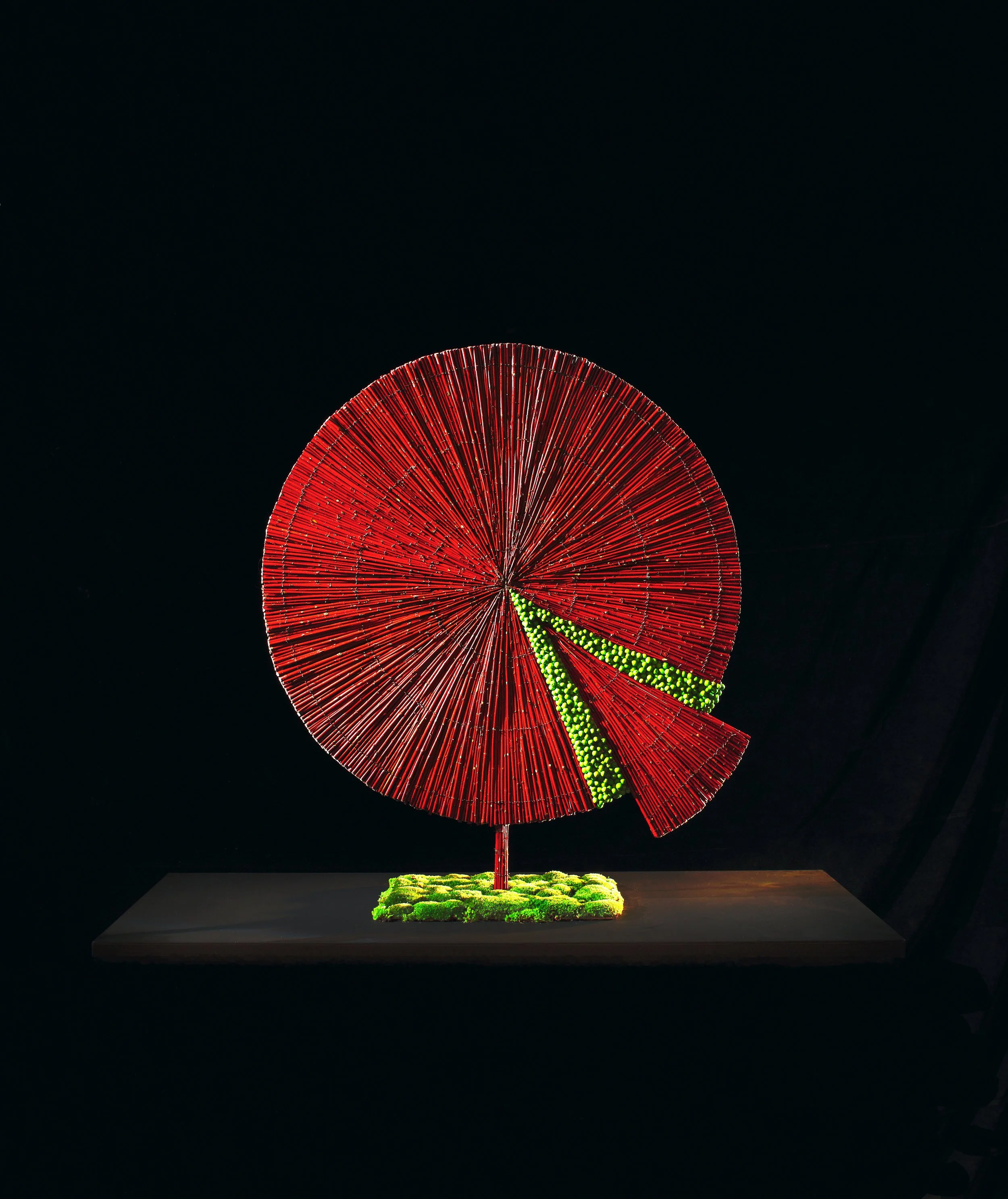 Architectural floral artwork shaped as a red circular fan with a geometric cut-out, decorated with vibrant green moss accents.