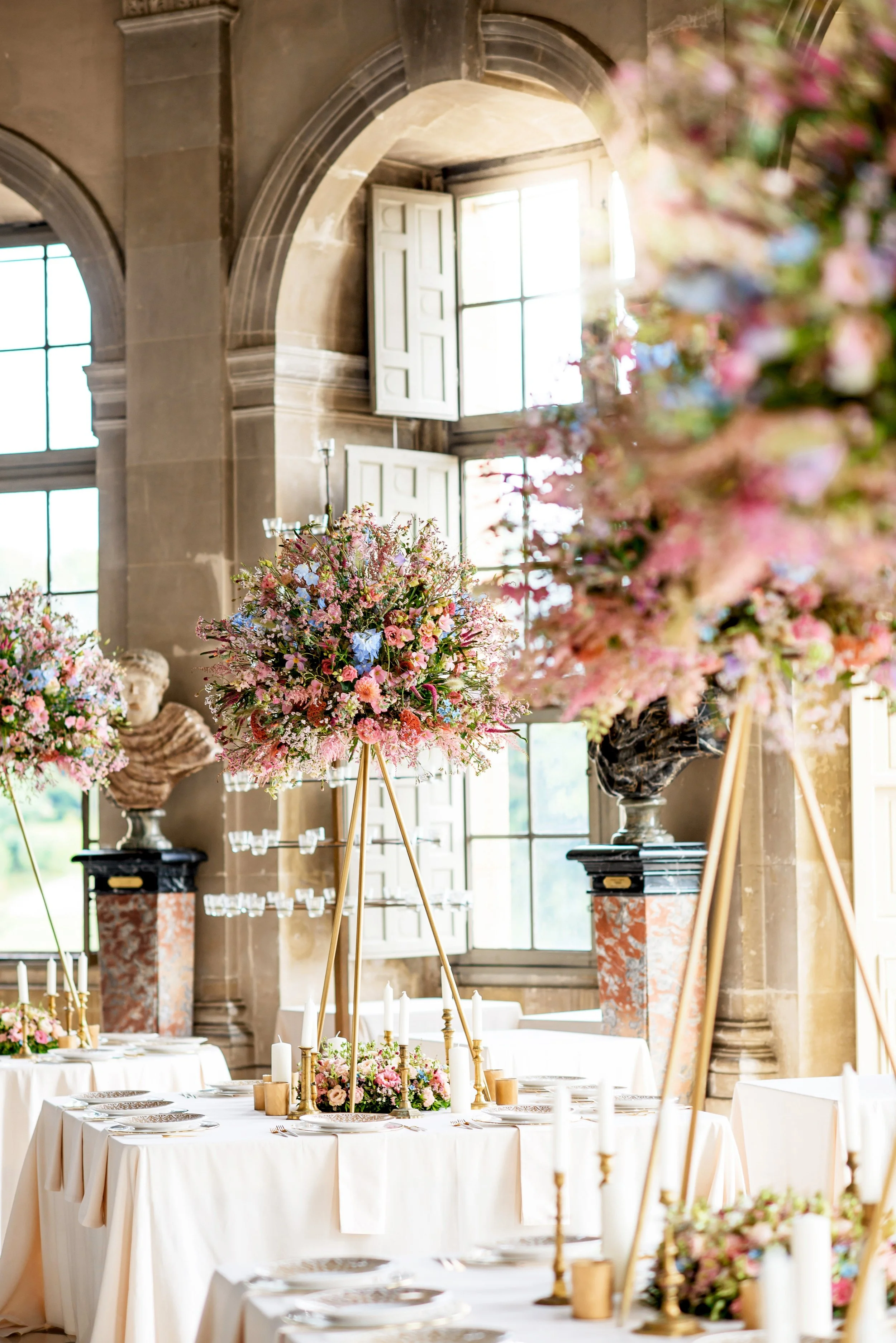 Romantic wedding table arrangement featuring tall golden tripod stands adorned with lush pink and blue blossoms, creating a luxurious and intimate dining atmosphere.