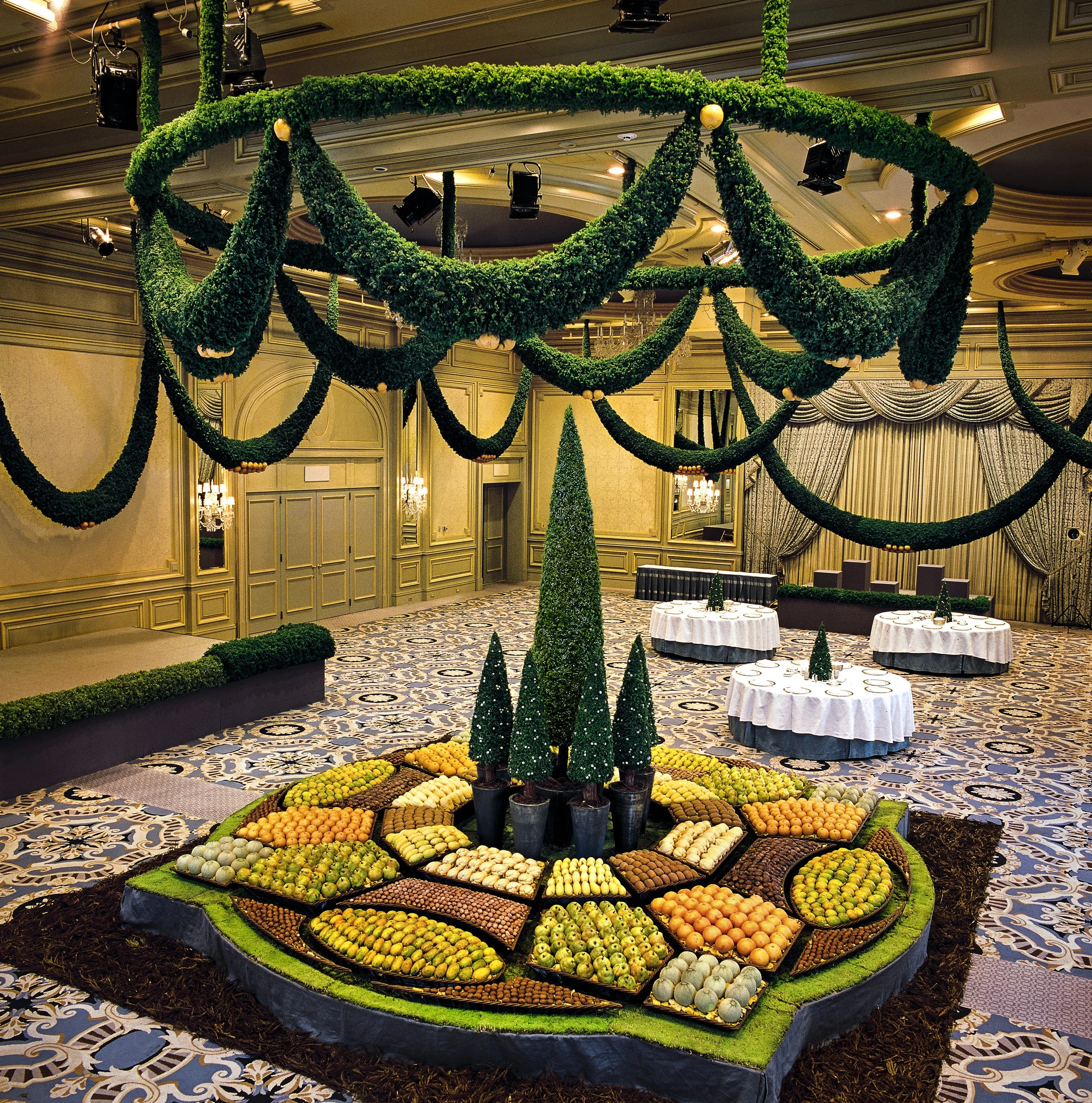 Grand ballroom installation with suspended greenery garlands, geometric fruit displays, and topiary trees arranged in a circular hospitality centerpiece.