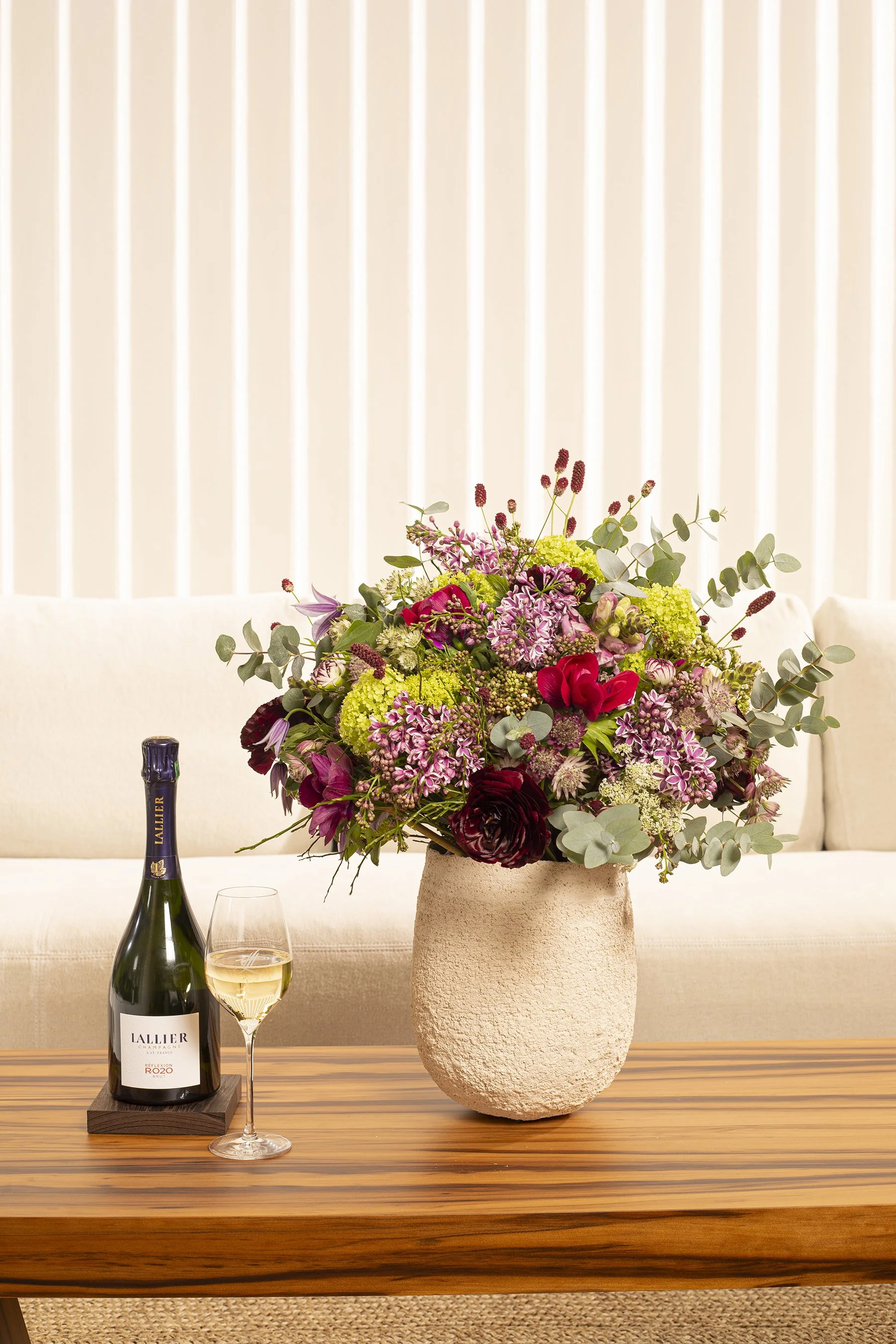 Elegant corporate reception bouquet in a textured stone vase, vibrant seasonal flowers with champagne, refined Daniel Ost atmosphere.