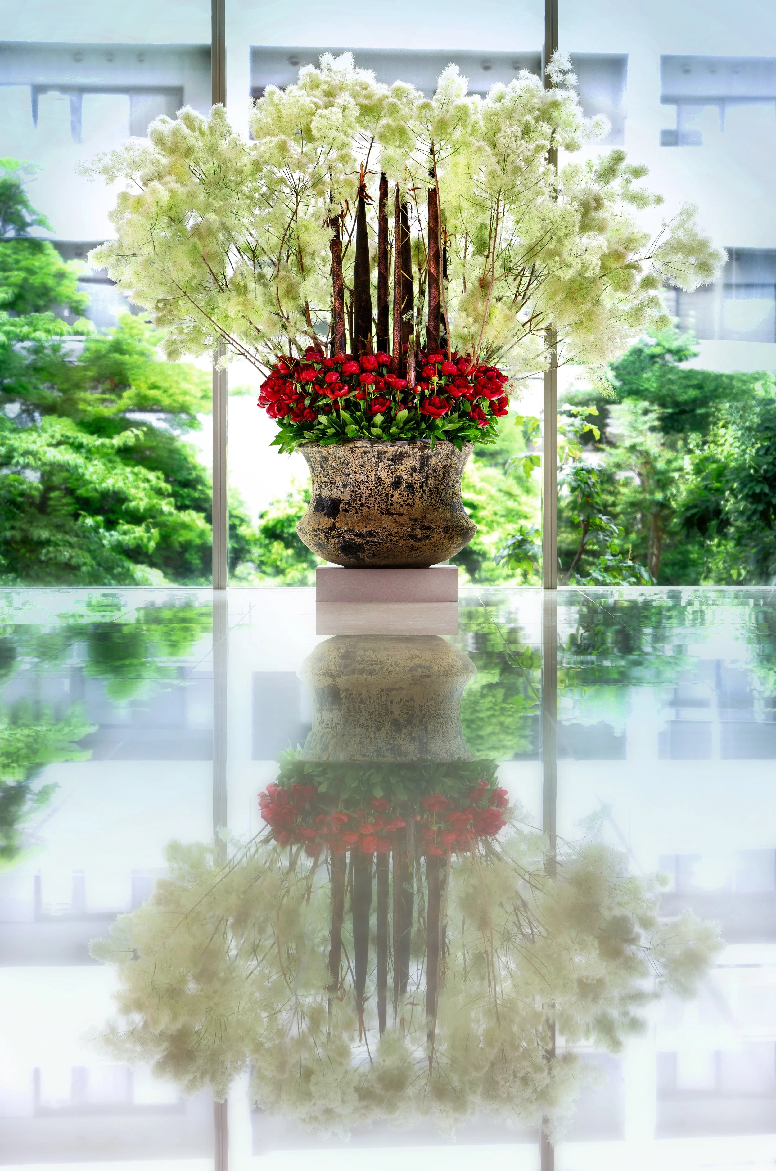 A large indoor floral arrangement with white fluffy flowers, red flowers, and green foliage in a rustic ceramic pot, with a mirror reflection on a shiny surface.