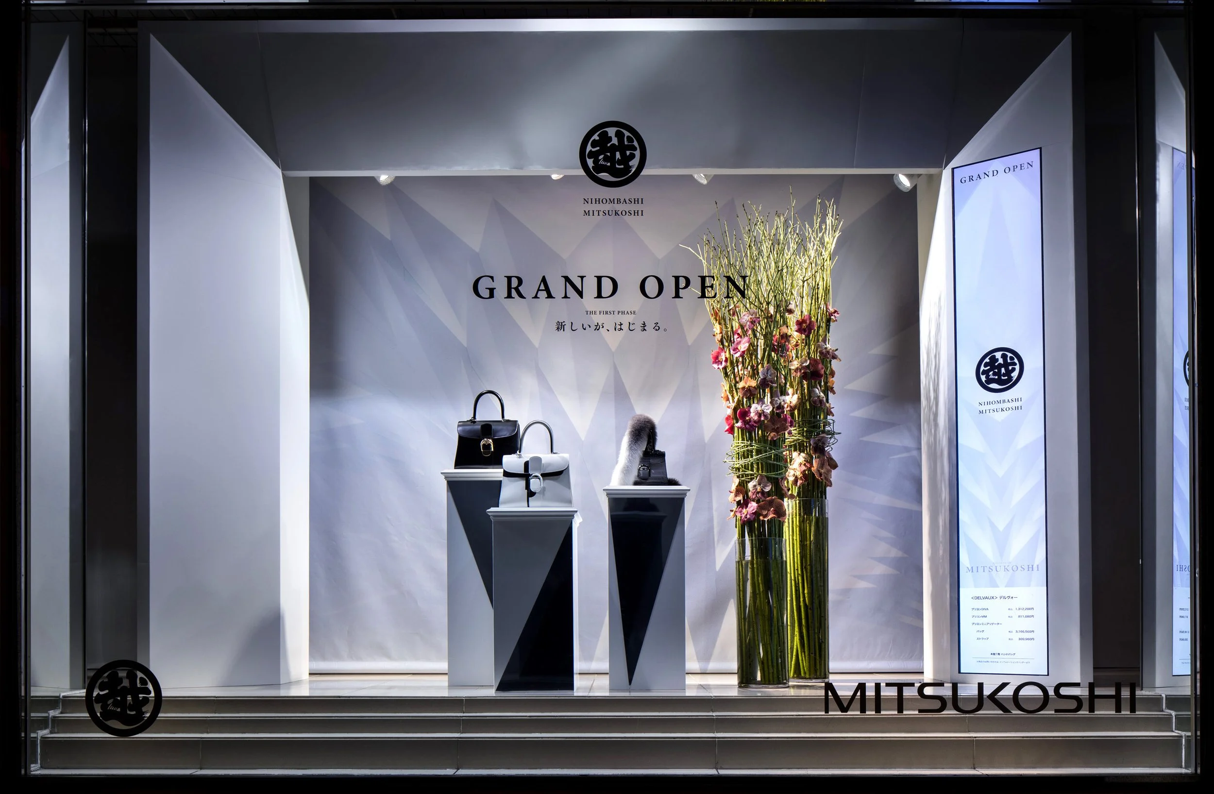 Store window display with three handbags on pedestals and tall vases with pink flowers, signage indicating grand opening of Mitsukoshi store.