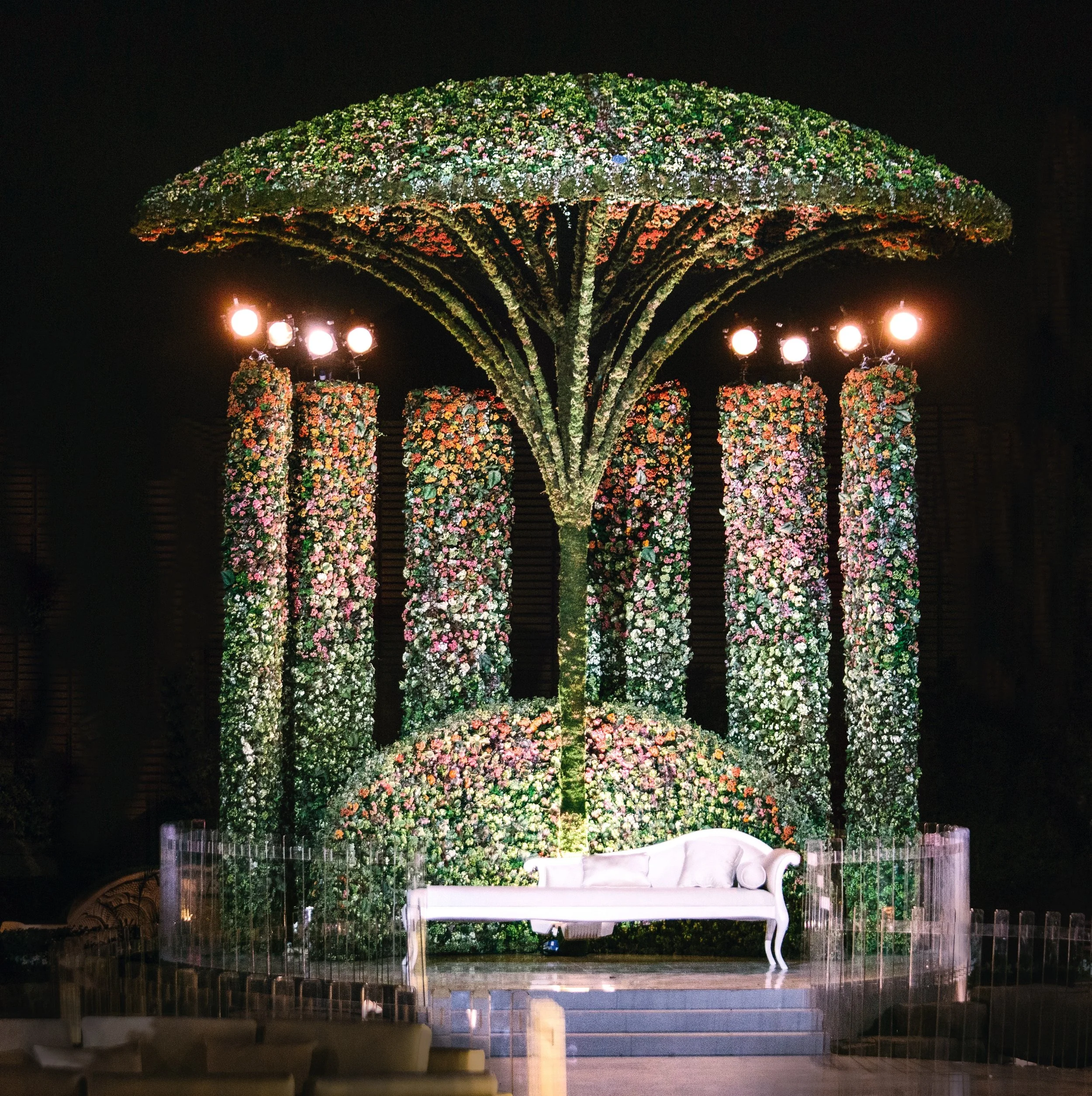 Dramatic outdoor wedding kosha stage framed by towering floral columns and a sculptural canopy of blossoms, with a white sofa centerpiece.