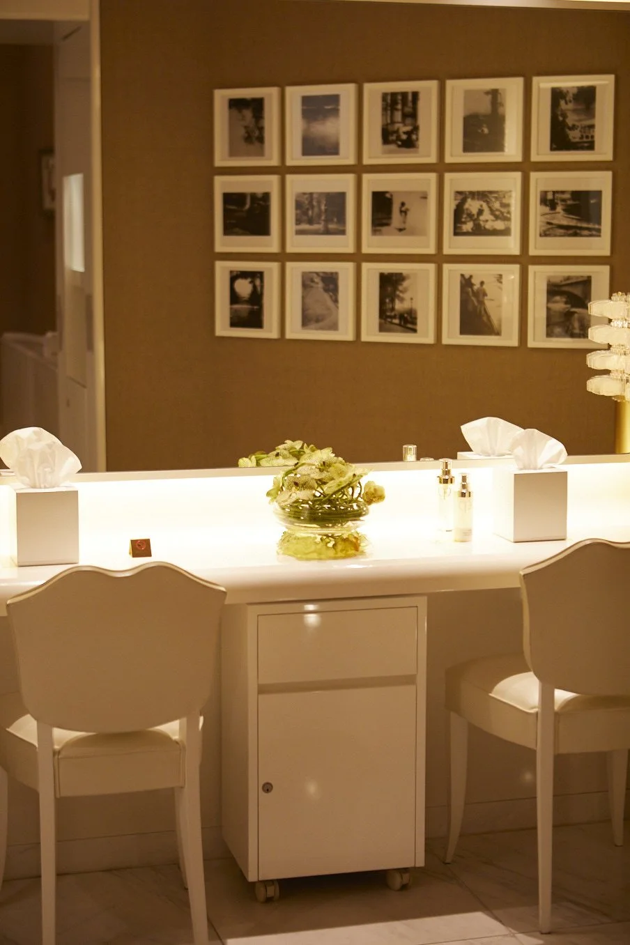 A well-lit bathroom vanity area with a backlit mirror, two white chairs, a small white cabinet, and a wall decorated with framed black-and-white photos.