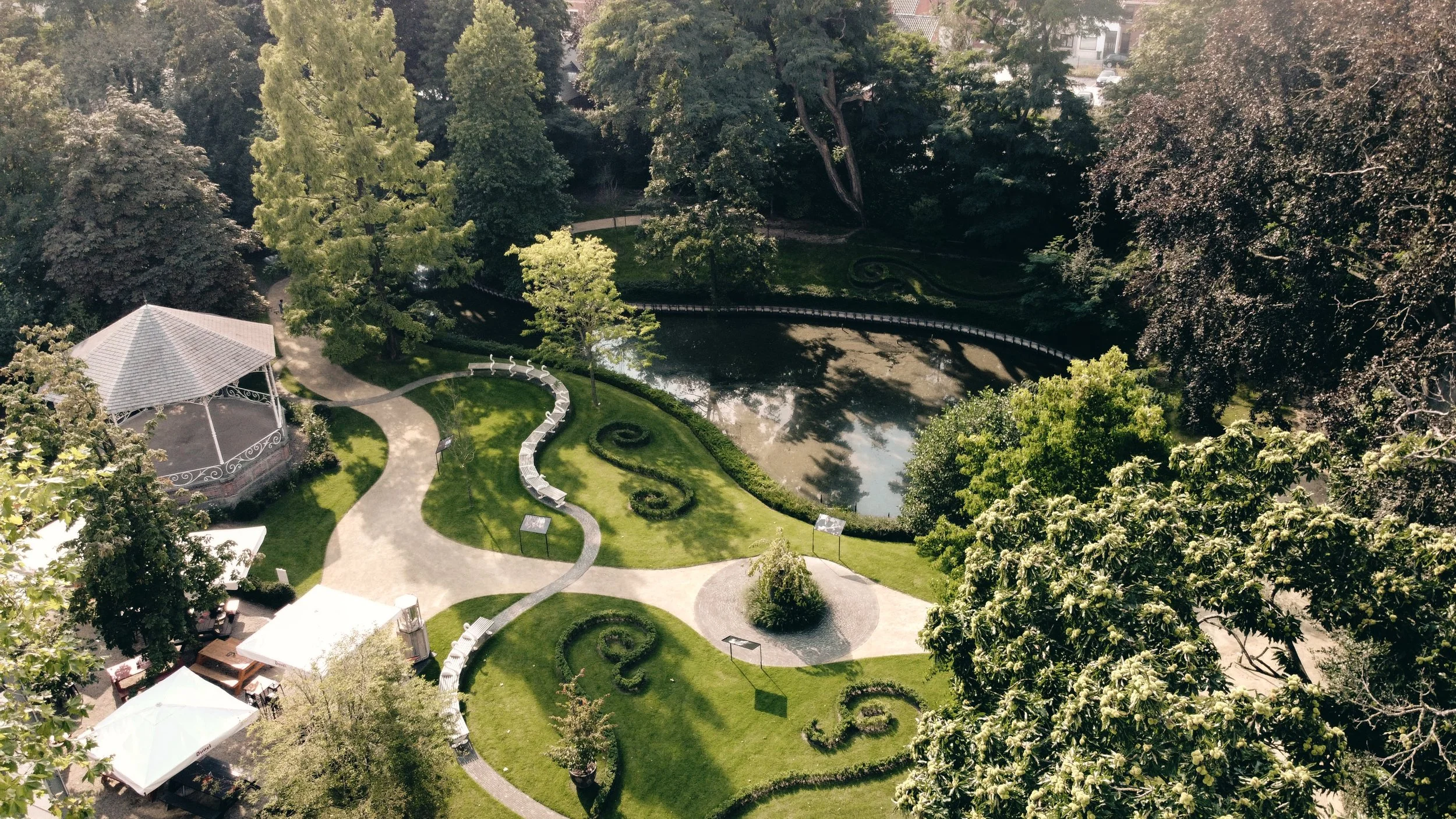 Aerial view of a landscaped park with a lake, winding pathways, and decorative garden patterns surrounded by mature trees.