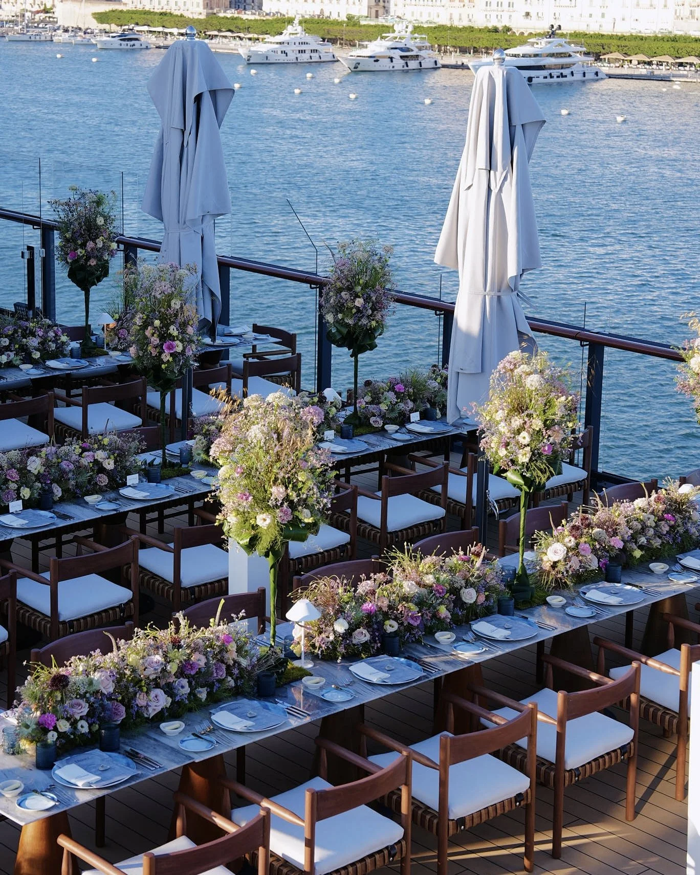 Outdoor dining setup on a deck overlooking a marina with boats, decorated with pink and purple flowers and white tableware, with closed white umbrellas.