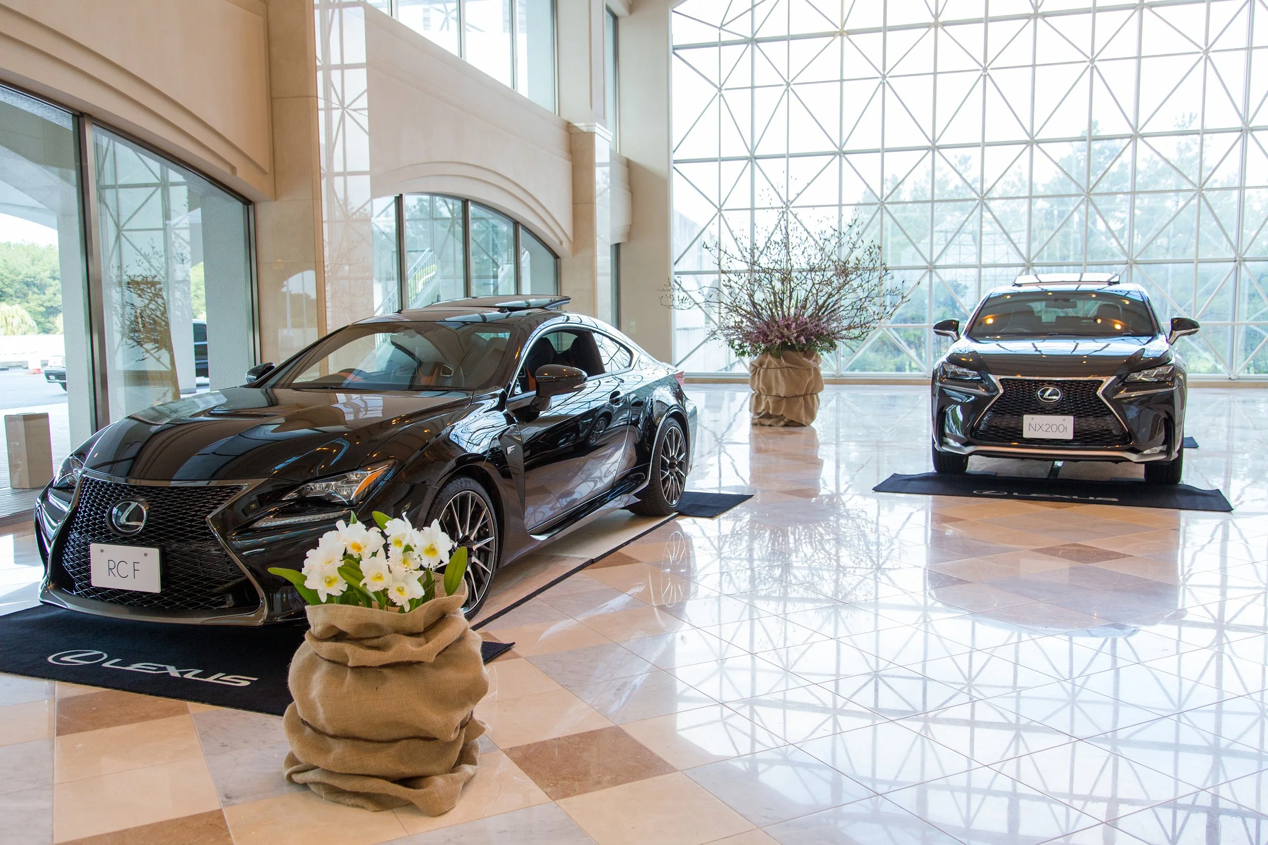 Corporate showroom display with Lexus cars framed by refined floral arrangements in neutral fabric planters, blending orchids and seasonal branches.