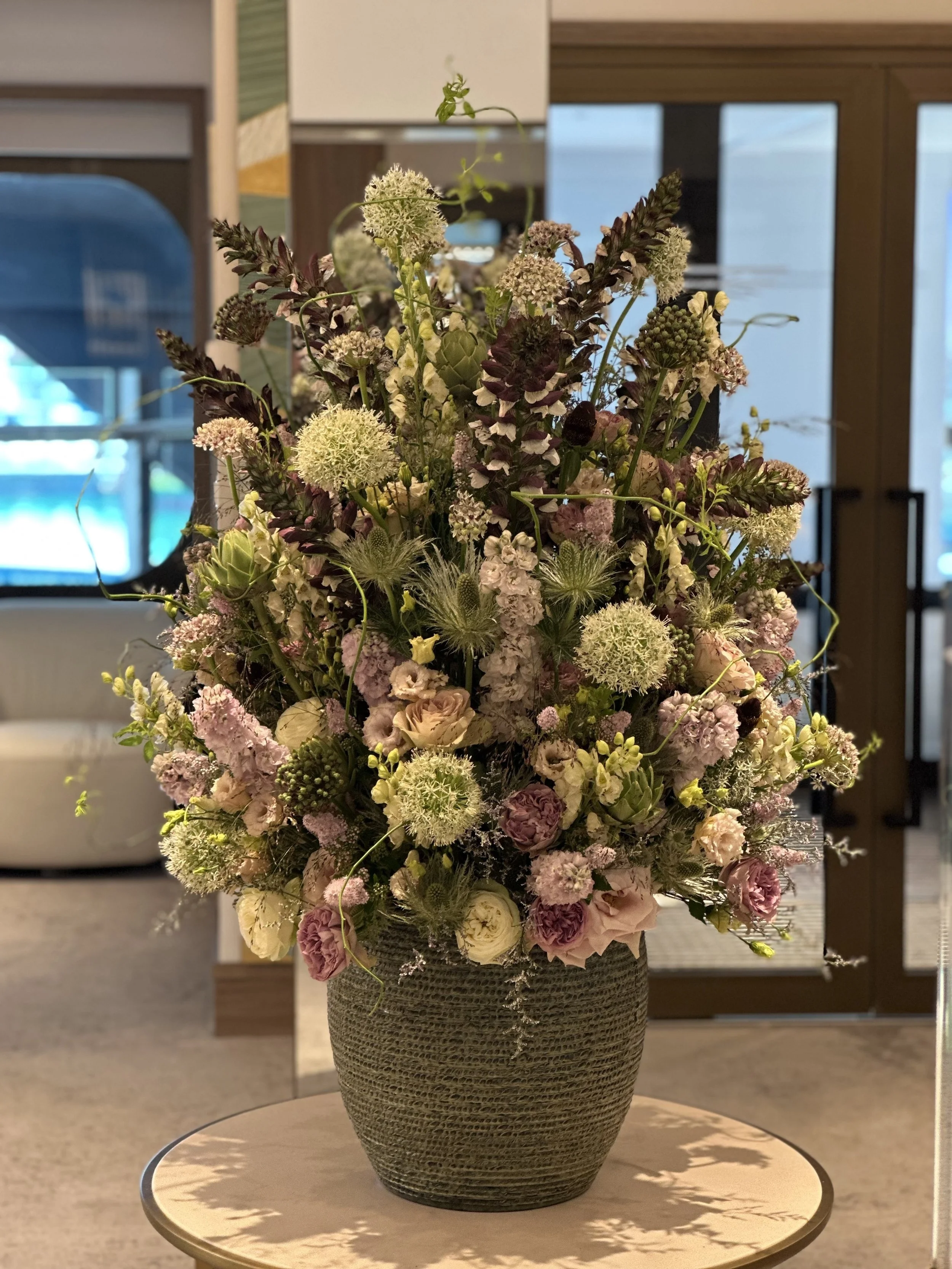 A large vase with a textured surface filled with a mixed arrangement of flowers and greenery, including white, pink, and purple blossoms, placed on a small round table indoors.