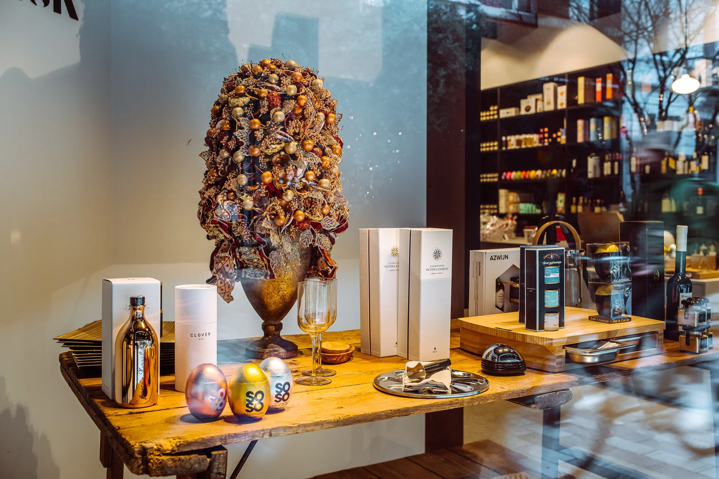 Display window with a decorated Christmas arrangement, including ornaments, a glass with a gold-colored drink, and various gift boxes and items on a wooden table inside the store.