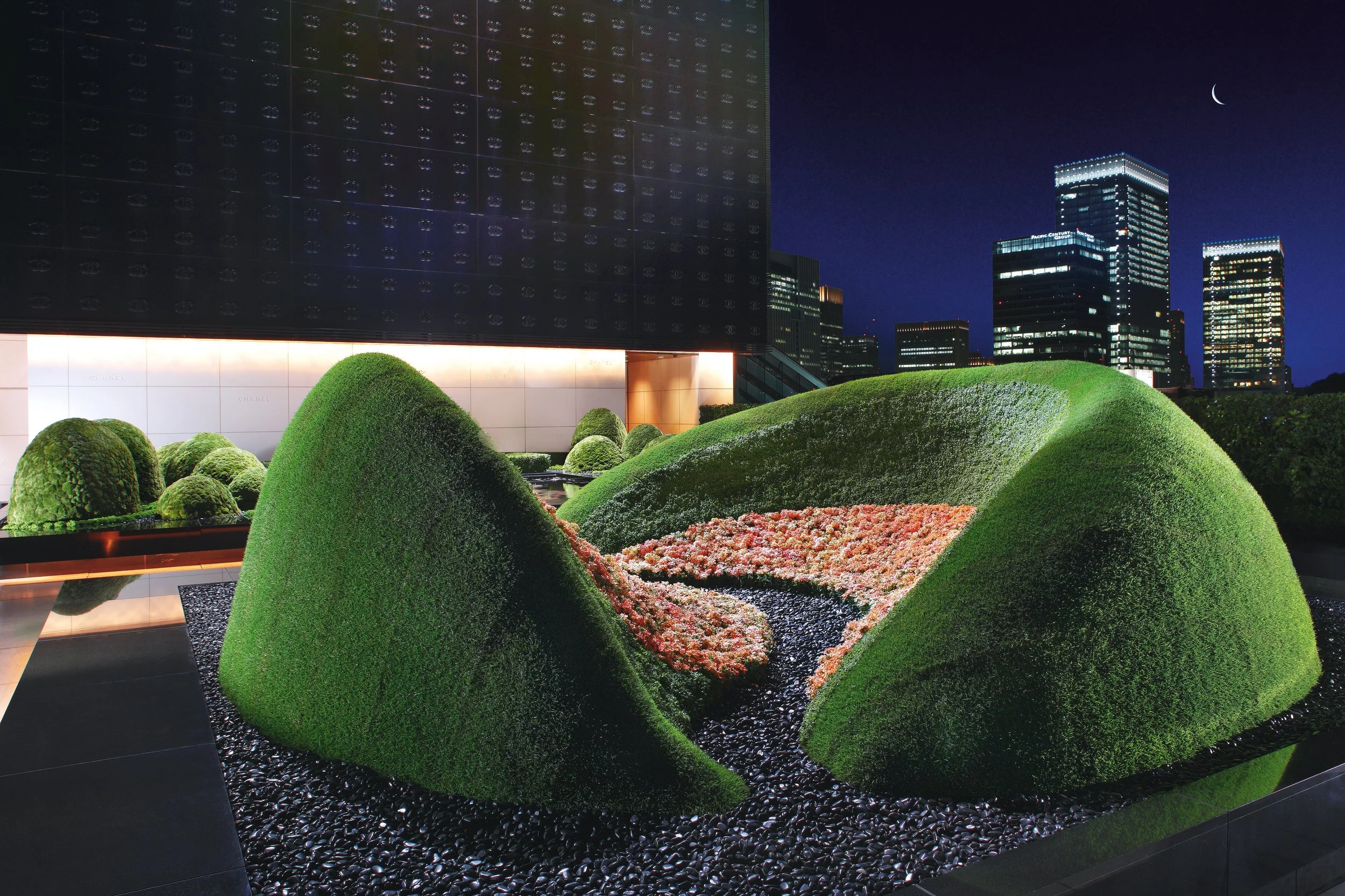Sculptural rooftop garden with abstract green mounds, floral river design, and a backdrop of illuminated skyscrapers at night.