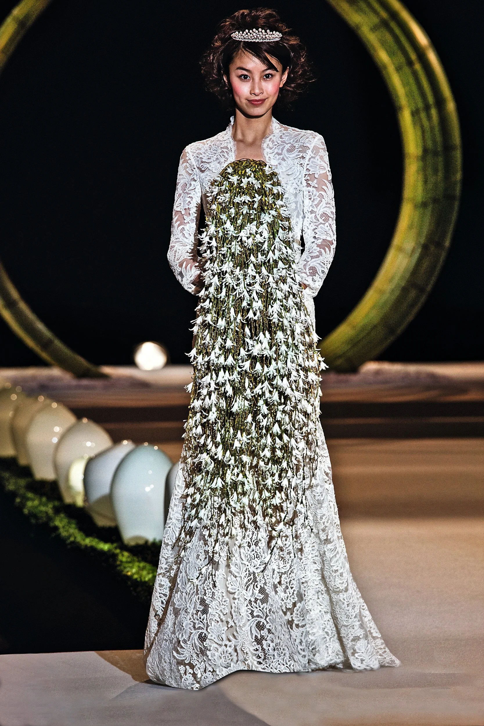A woman wearing a white lace dress with intricate floral details and a tiara, standing on a runway with a large circular backdrop behind her.