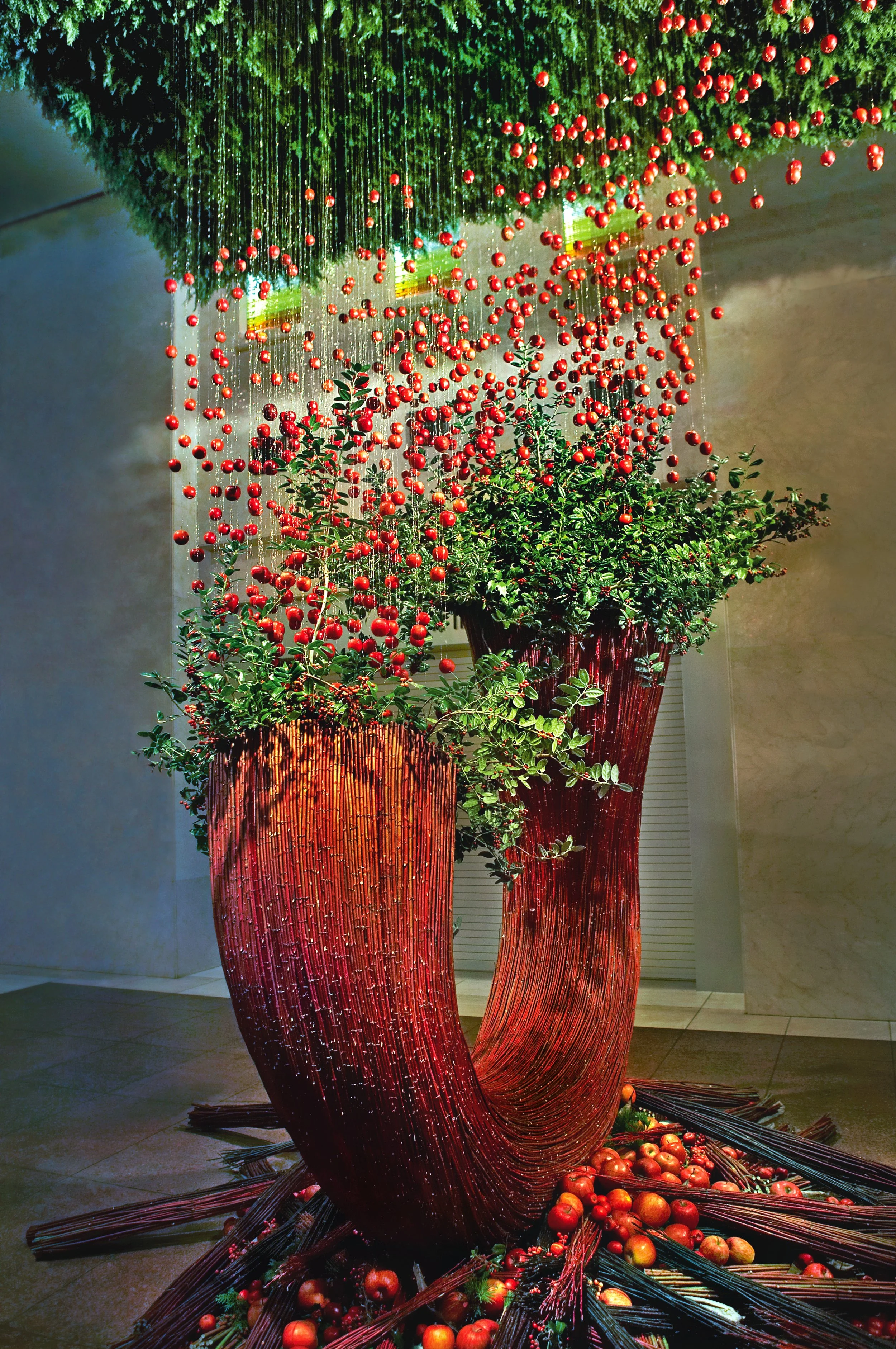 Large sculptural floral installation with red bamboo arcs, green foliage, and cascading suspended apples, styled for a luxury hospitality lobby.