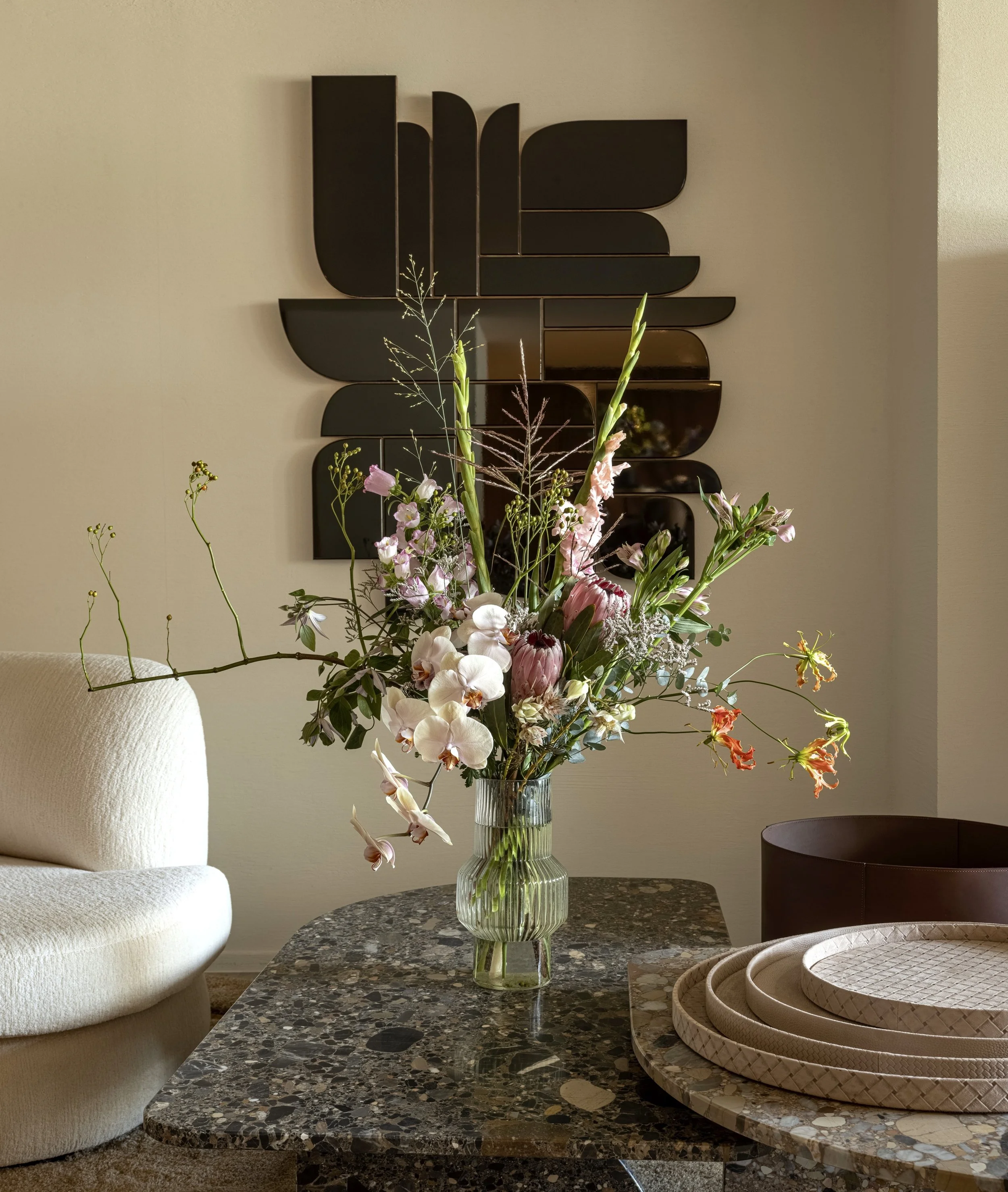 Decorative living room scene with a tall glass vase of pink and white flowers on a dark granite table, a beige armchair, black wall art, and layered round trays on the right.