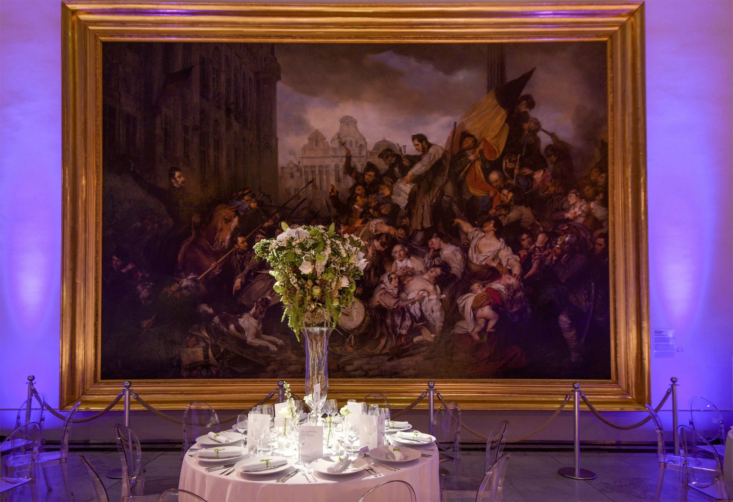 Wedding dining table set beneath a dramatic historic painting, featuring a tall glass vase centerpiece with white flowers and greenery for a refined atmosphere.