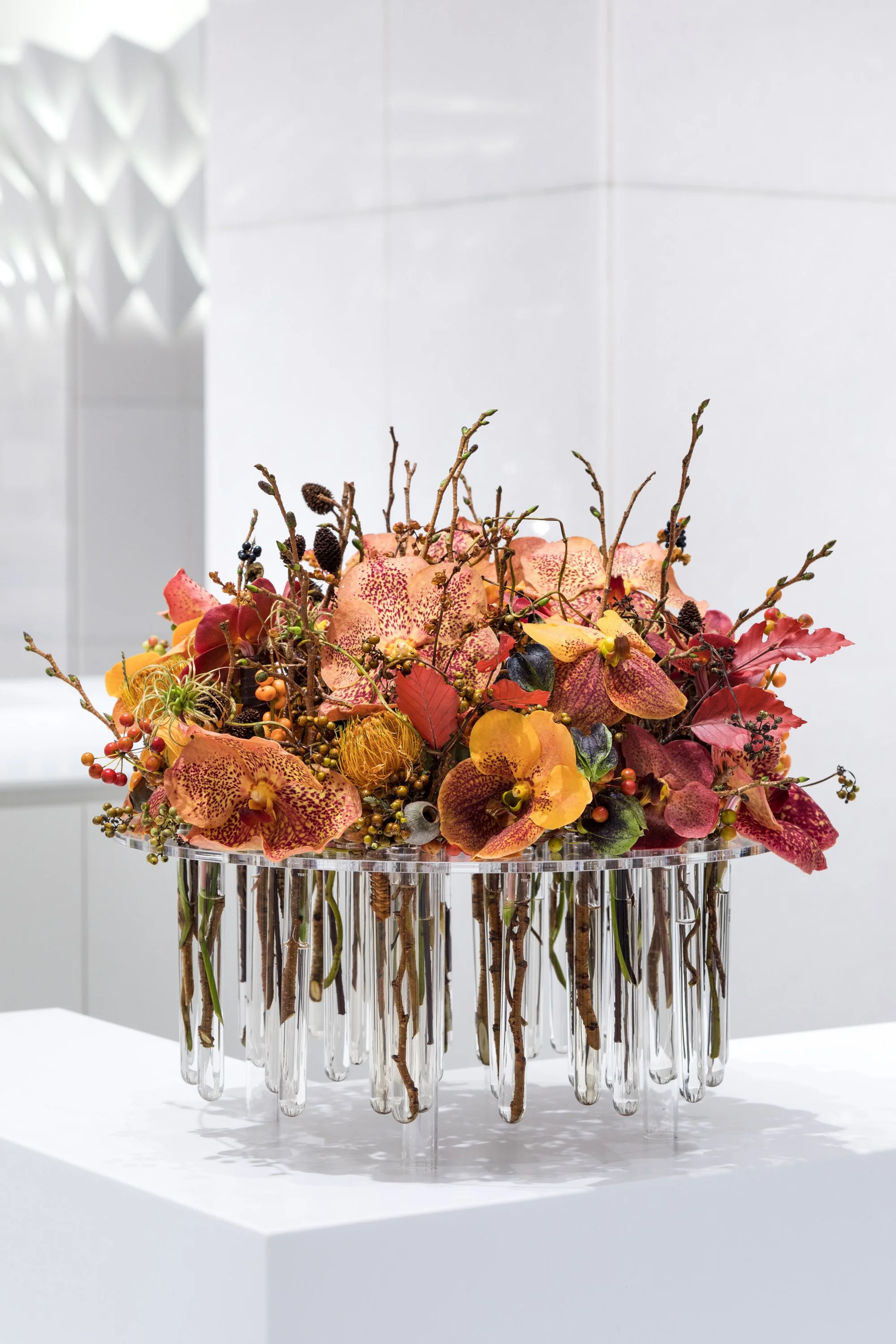 Autumn floral arrangement with orange and yellow flowers, branches, berries, and foliage displayed in a glass vase with hanging glass tubes, on a white pedestal.