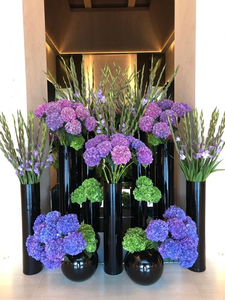 Hotel lobby floral display with layered purple hydrangeas, green hydrangeas, and gladiolus in glossy black vases.