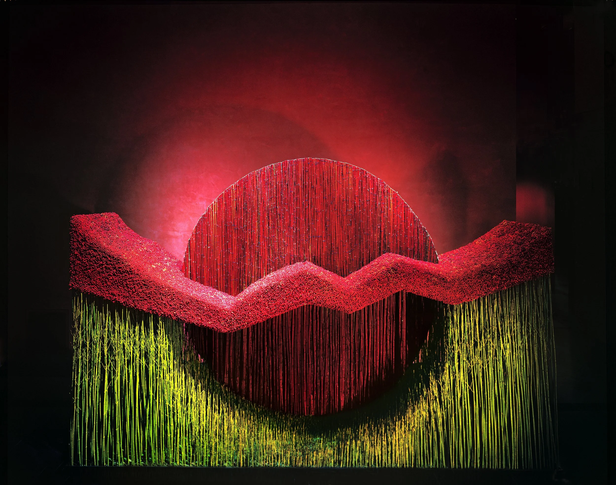 A modern art installation featuring a large red semi-sphere with vertical lines, surrounded by pink and yellow vertical rods, set against a dark background with red light effects.