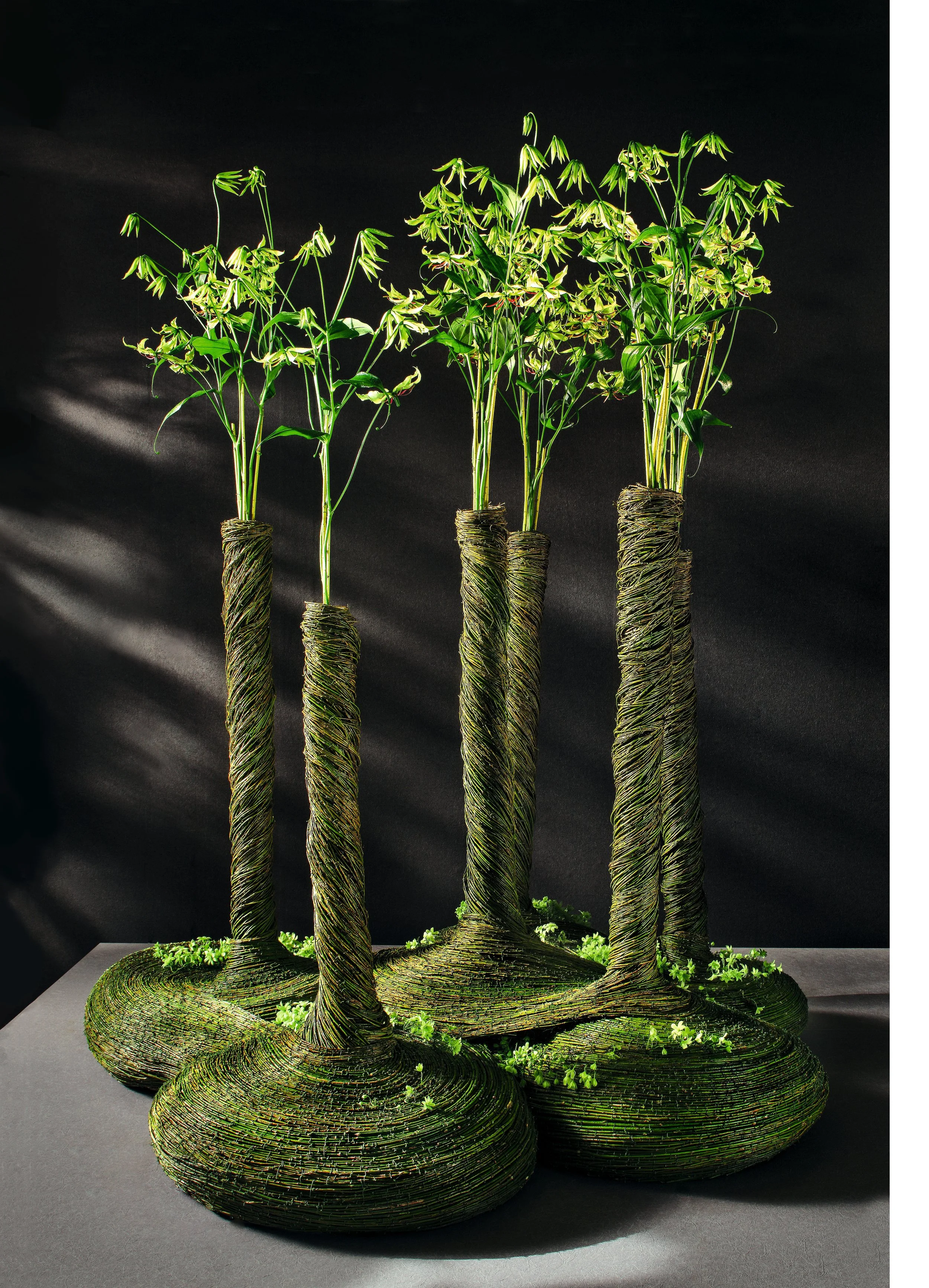 Clustered sculptural floral arrangement with tall stems wrapped in moss, topped with green blossoms, resembling miniature tree columns.