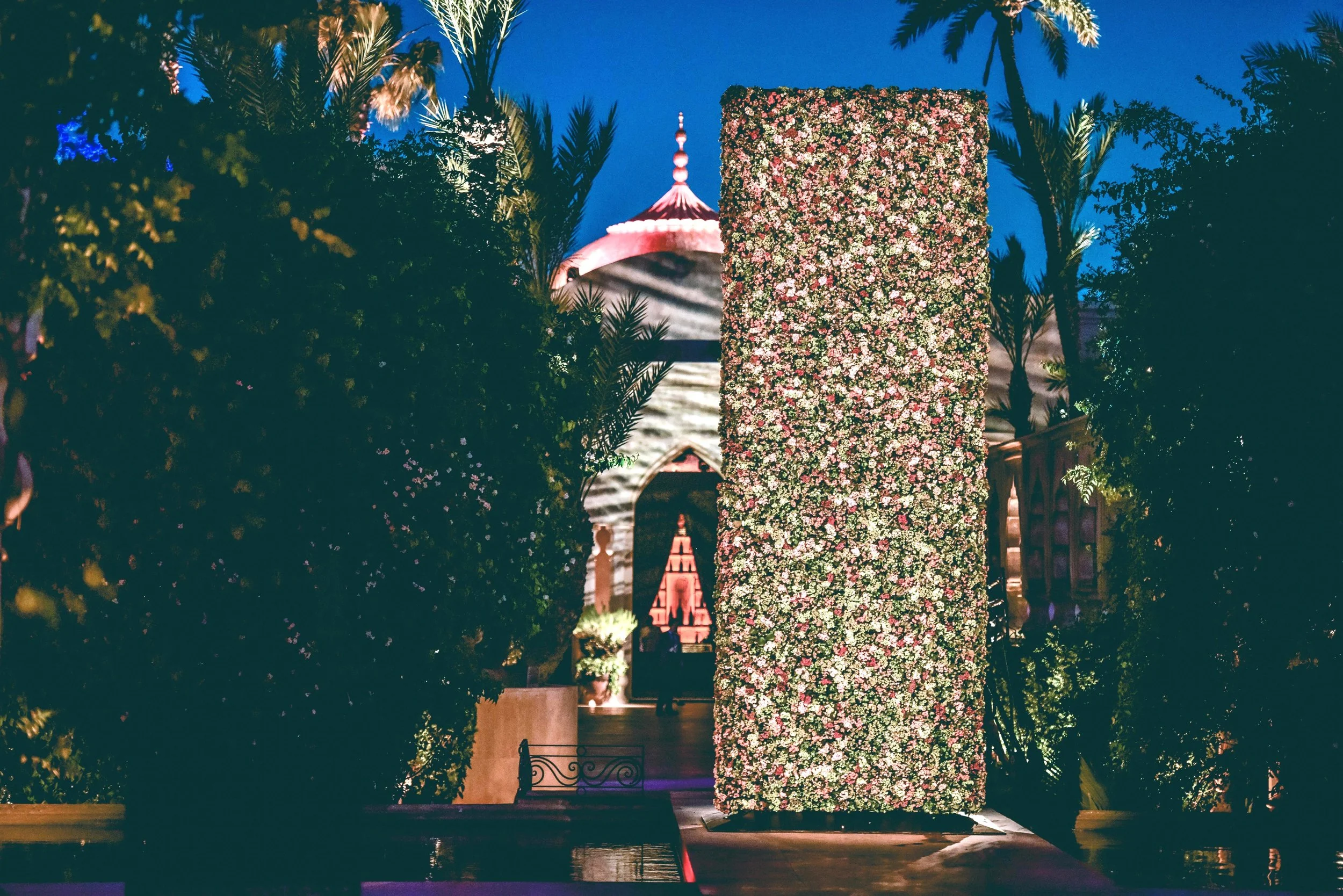 Outdoor luxury wedding in Marrakesh with a dramatic floral wall installation surrounded by palm trees and palace architecture.