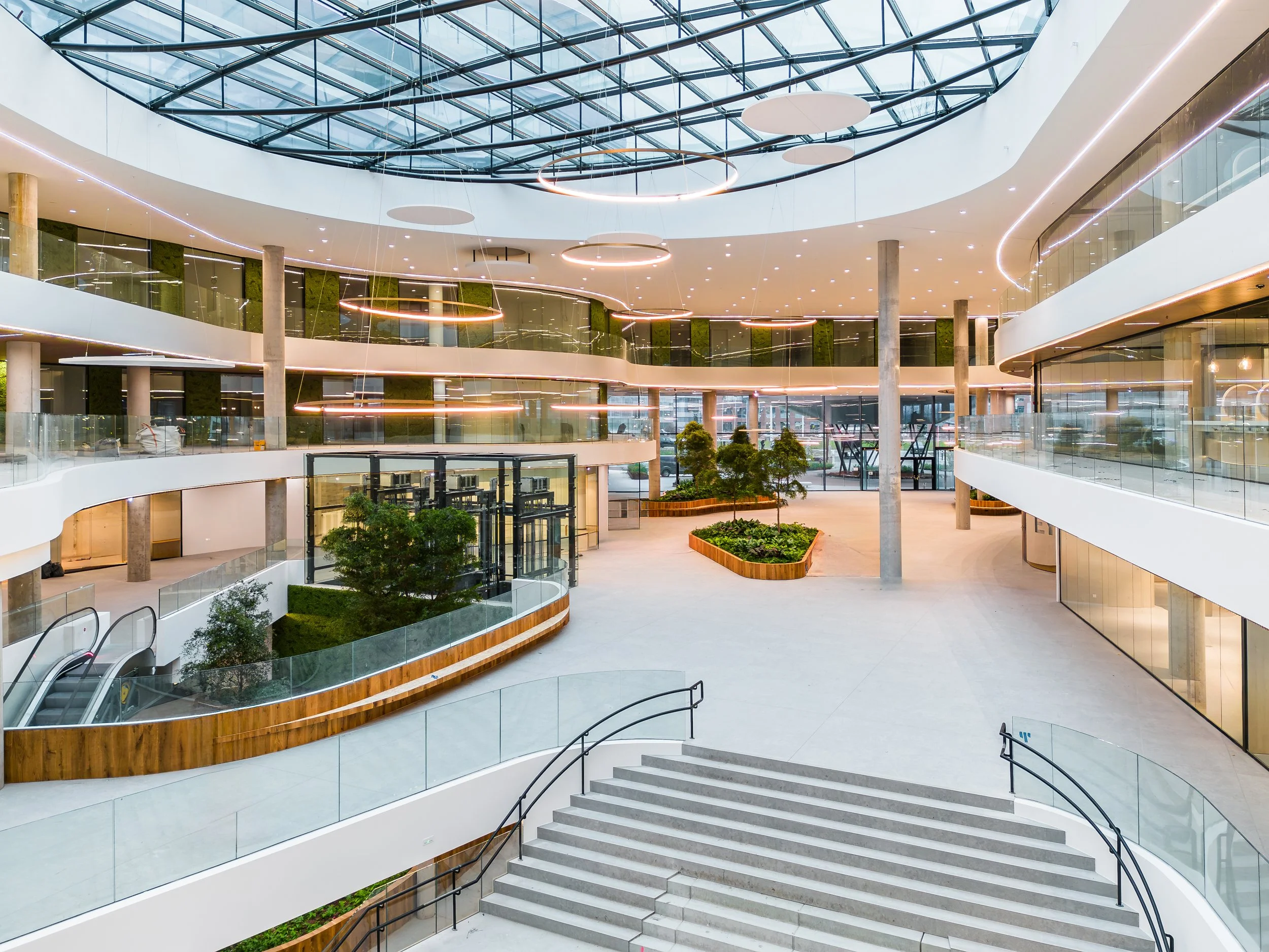 Modern office atrium with skylight ceiling, indoor greenery, wide staircases, and open-plan terraces with glass railings.