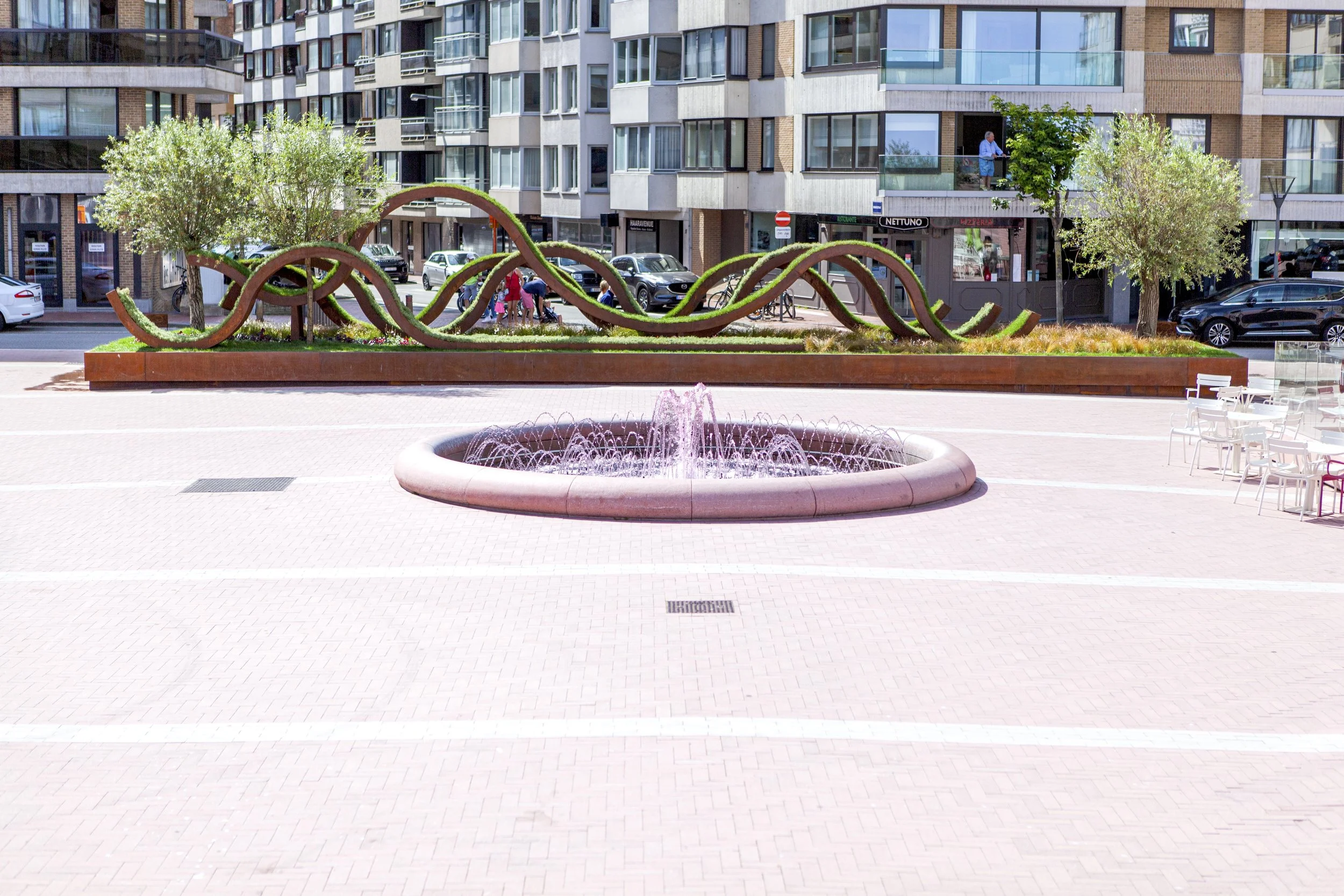 Urban plaza with pink fountain, olive trees, and a contemporary sculptural installation of looping grass forms.