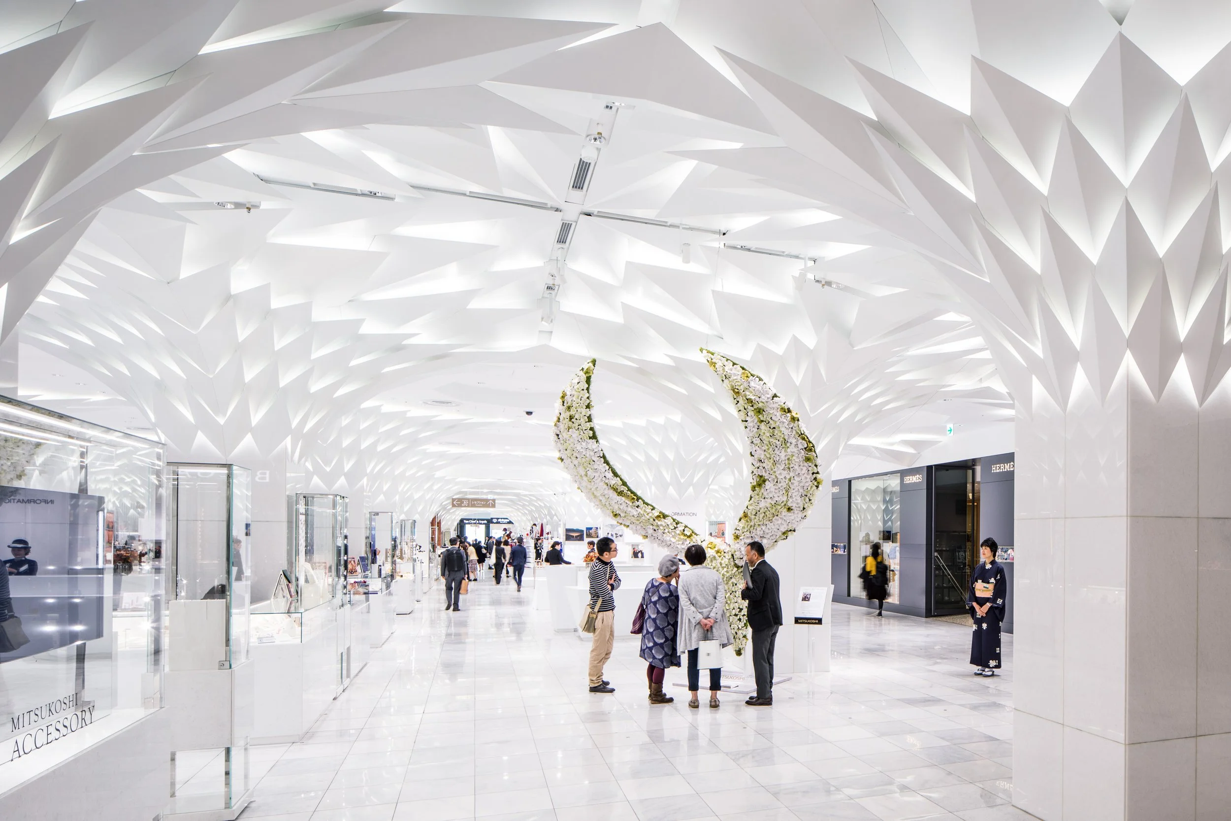 Modern, brightly lit airport terminal with white geometric ceiling design, floral sculpture in the center, and travelers walking or waiting.
