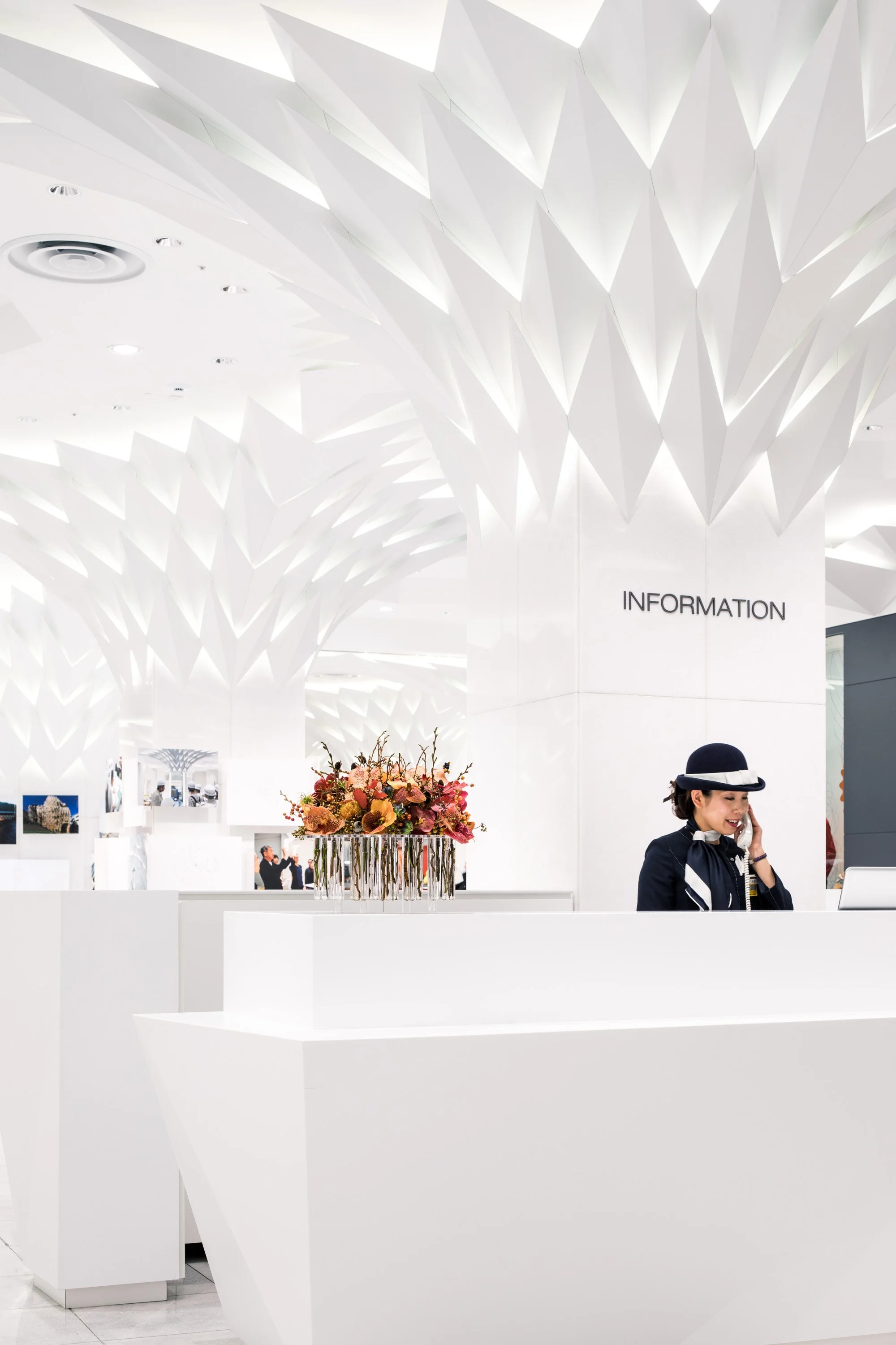 Interior of a modern, white information desk with a floral arrangement, and a woman talking on the phone. Signage reads 'INFORMATION' on the wall.